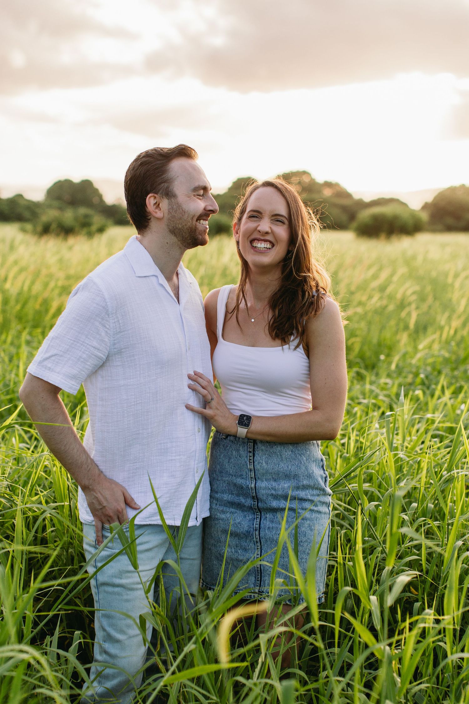 Happy couple laughing together while standing in a lush green field at sunset.