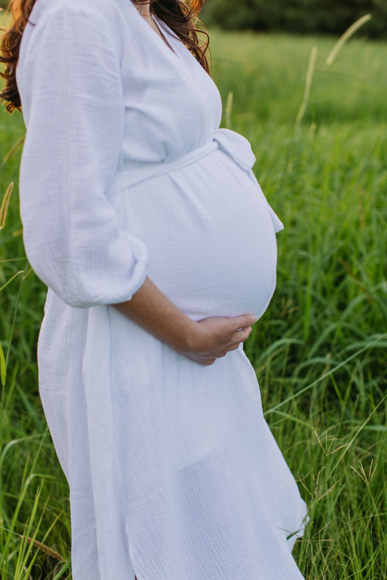 Pregnant woman in white dress cradling baby bump in lush green field.