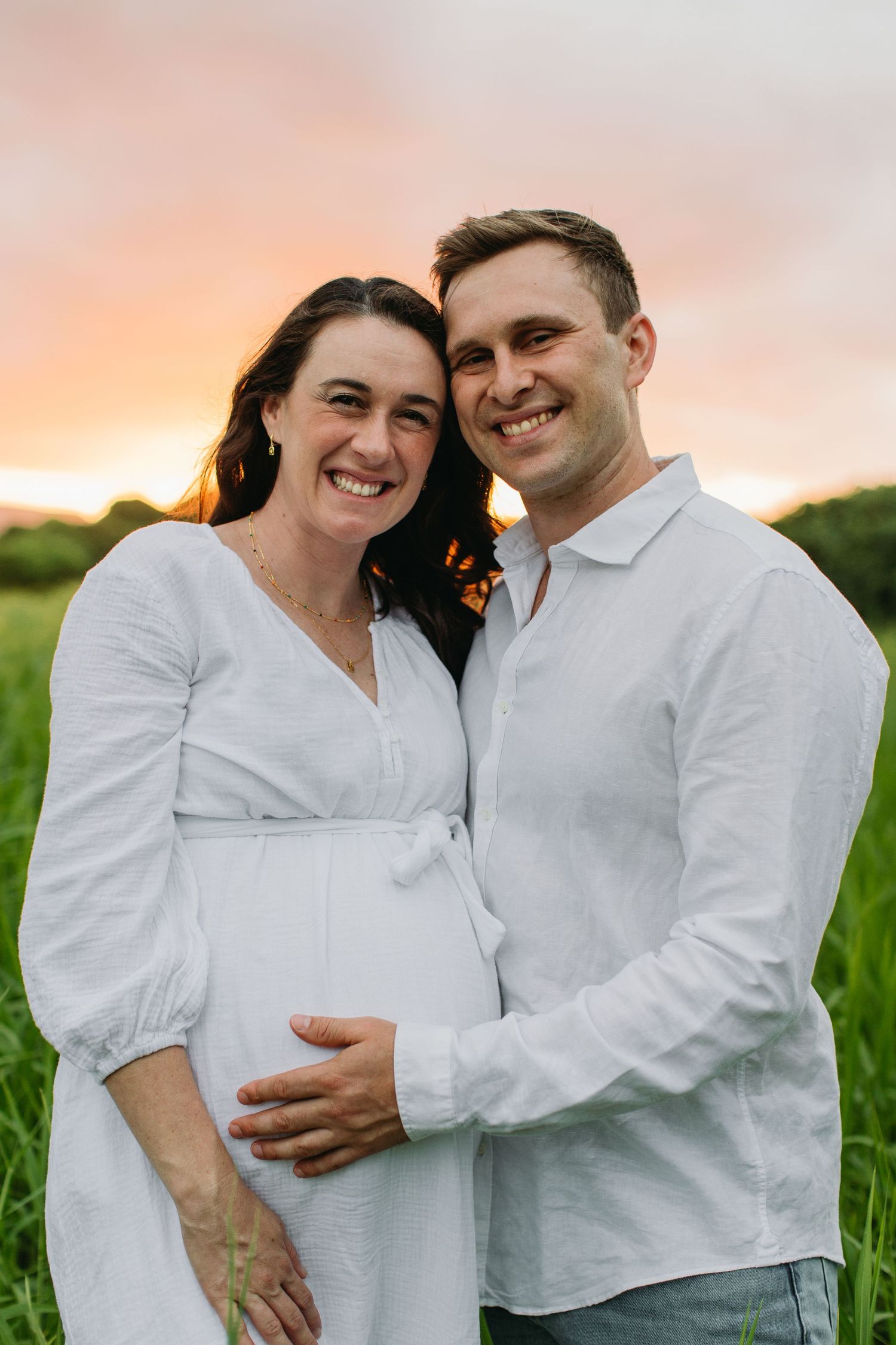 Smiling pregnant couple in white outfits pose in a green field at sunset, man gently holding woman's baby bump.