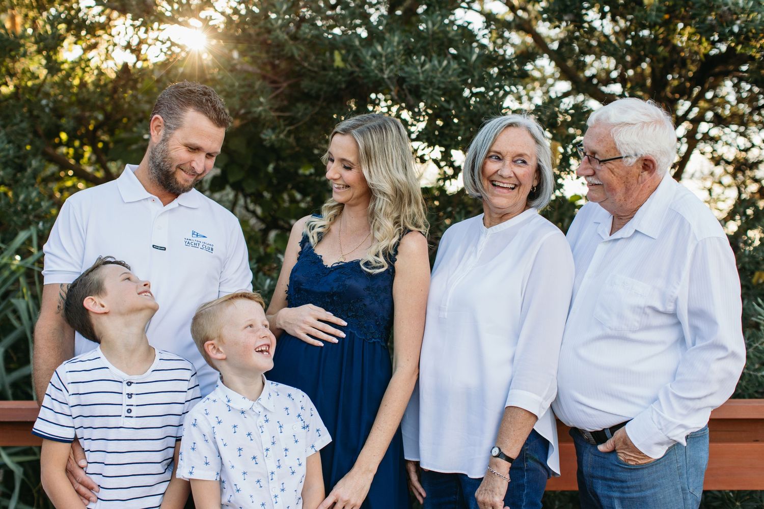 A happy multigenerational family poses outdoors, with a pregnant woman at center, two boys, a couple, and grandparents smiling.