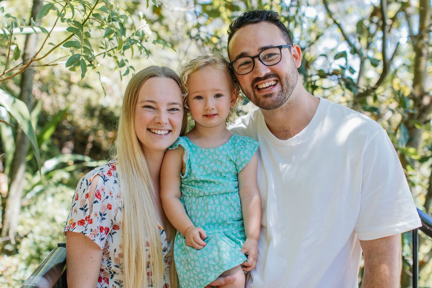 Happy family portrait of smiling parents holding their young daughter outdoors in a lush green garden setting.