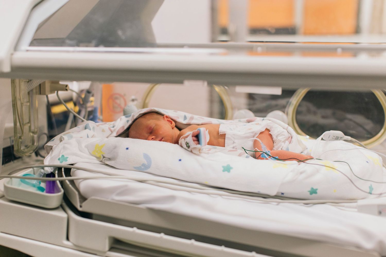 Premature newborn baby resting in a hospital incubator wrapped in a white blanket with colorful star pattern.