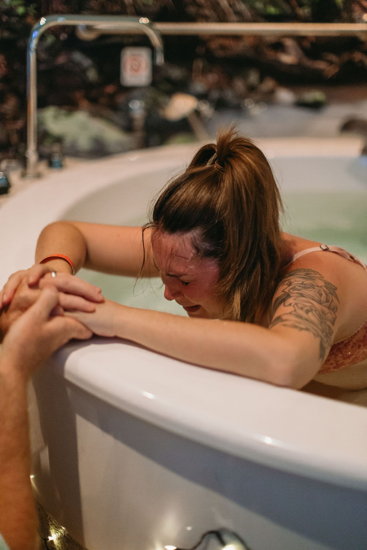 Woman in bikini leaning over edge of birthing tub, holding hands with support person during water birth labor.