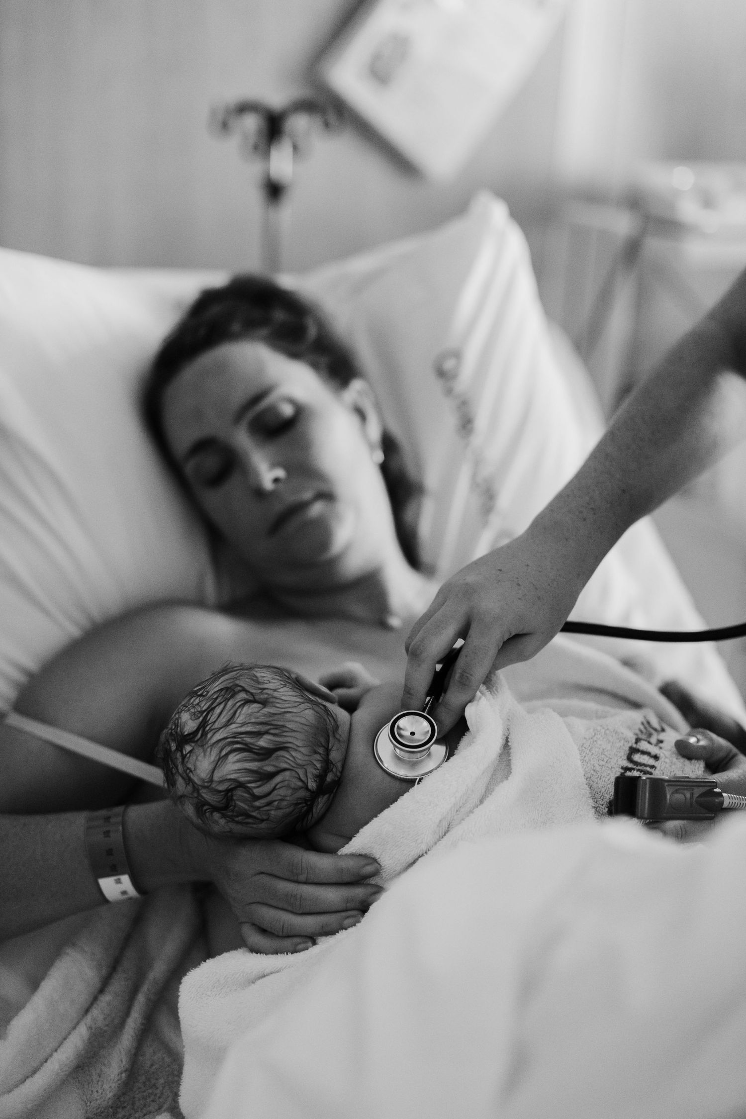 New mother lying in hospital bed holding newborn while nurse checks baby with stethoscope, black and white photo.