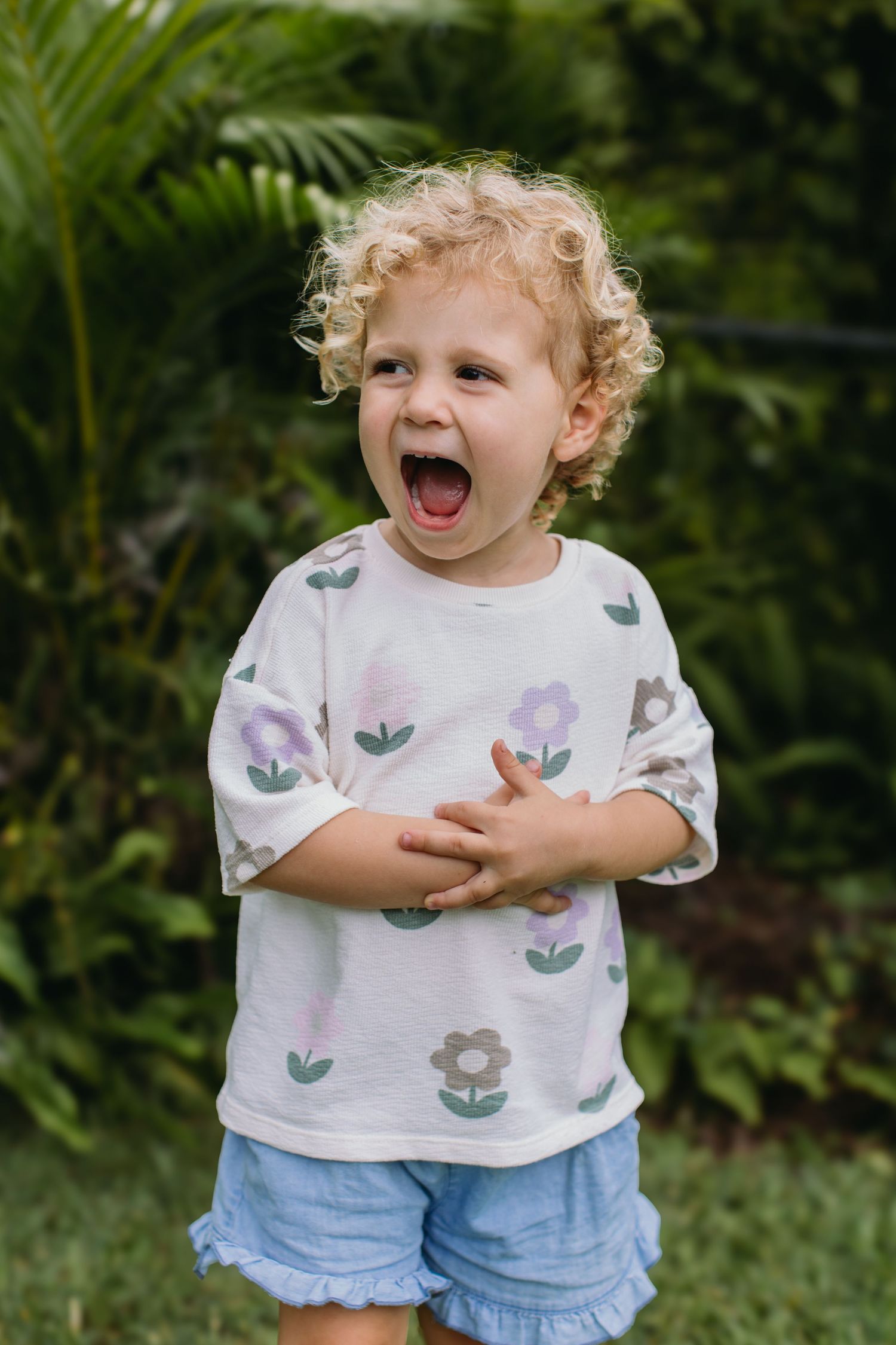 Curly-haired toddler in floral shirt and blue shorts laughing outdoors in a lush green garden.