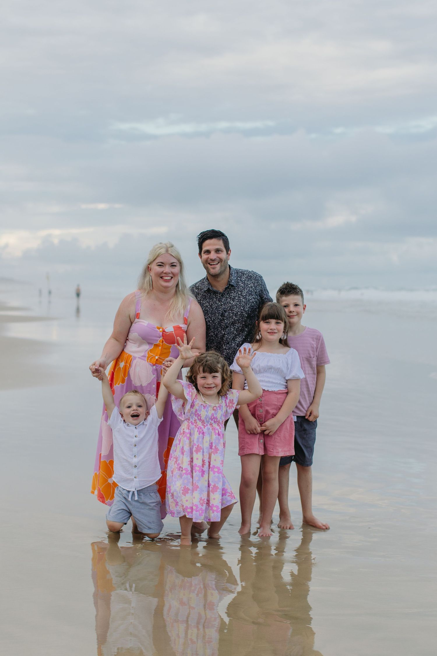 Happy family of six standing in shallow ocean water on a cloudy beach day, children waving.