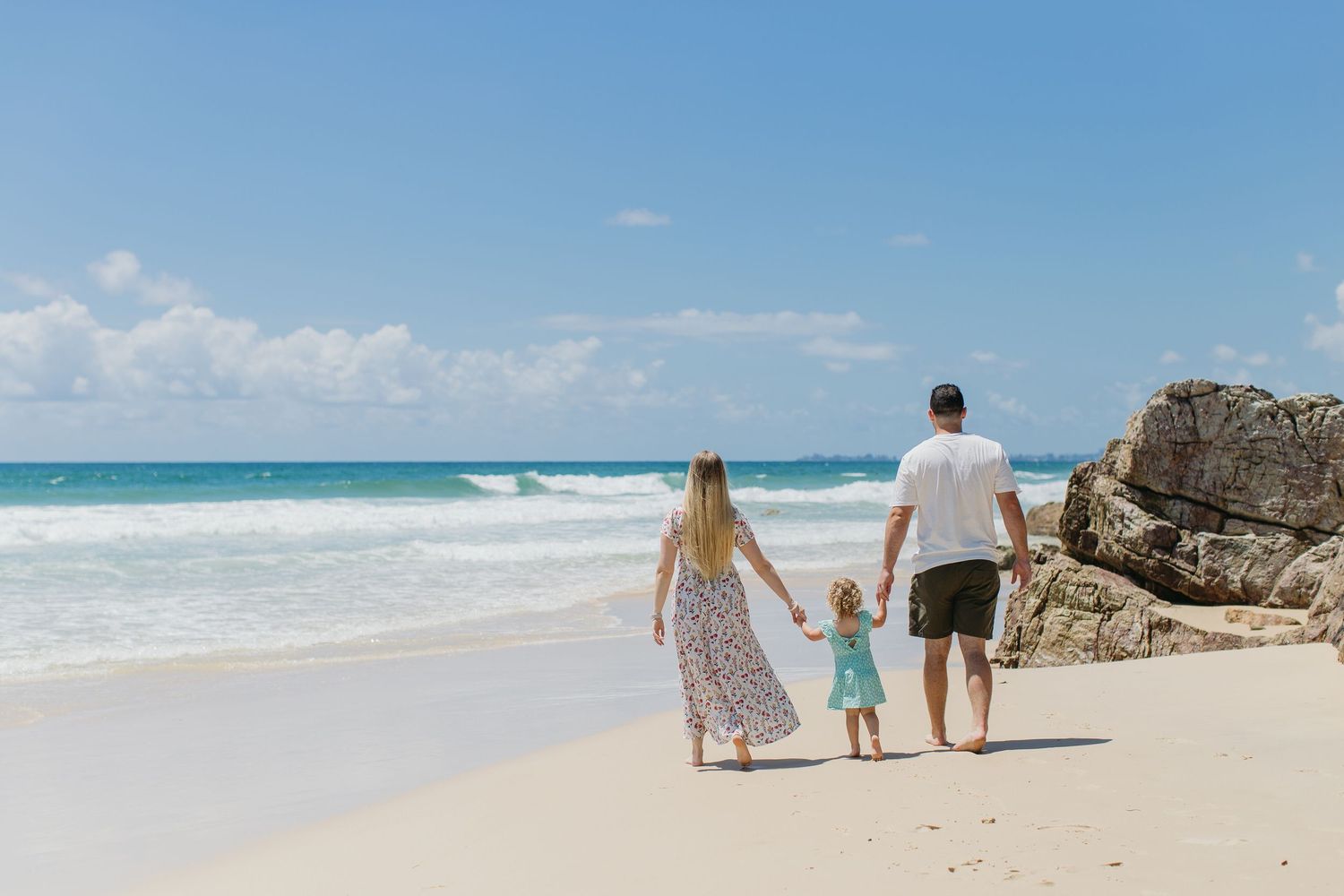 Family of three holding hands walking on a sunny white sand beach with turquoise ocean waves and rocky cliffs.