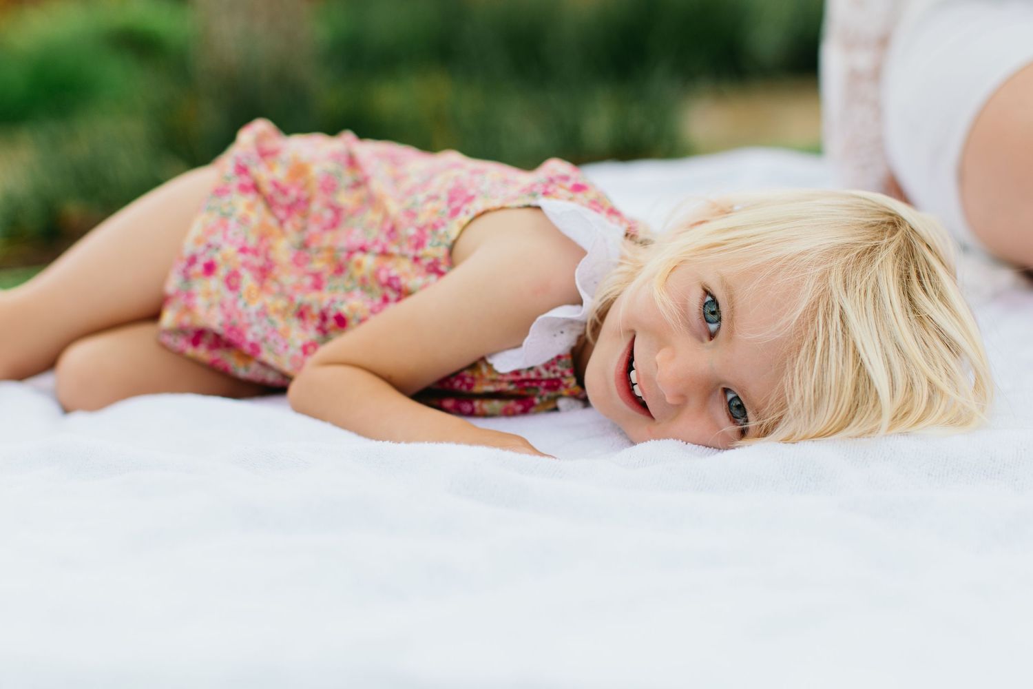 Smiling blonde toddler girl in floral romper lying on white blanket outdoors.