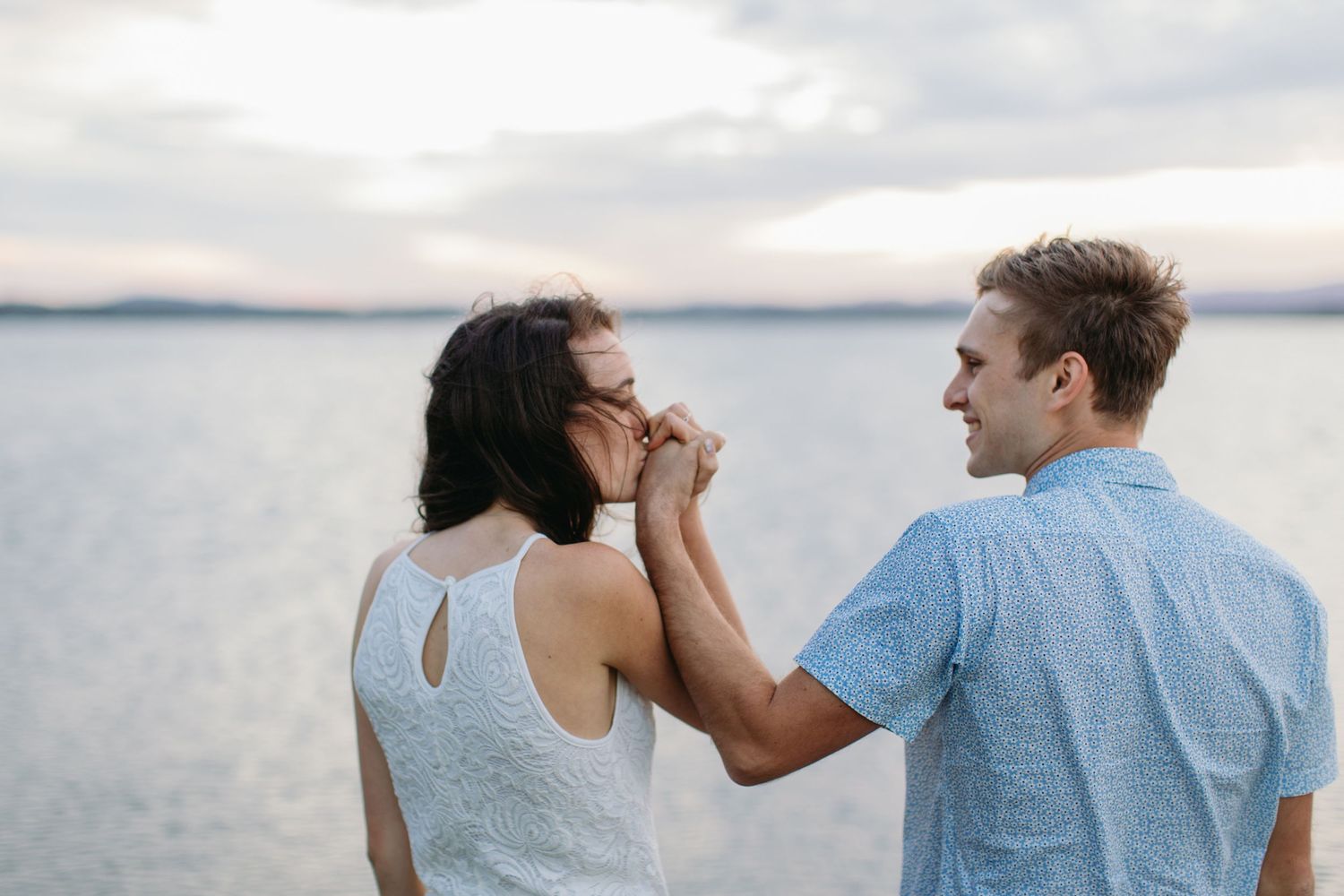 Romantic couple holding hands by the lake at sunset, man smiling at woman in white dress near calm water.