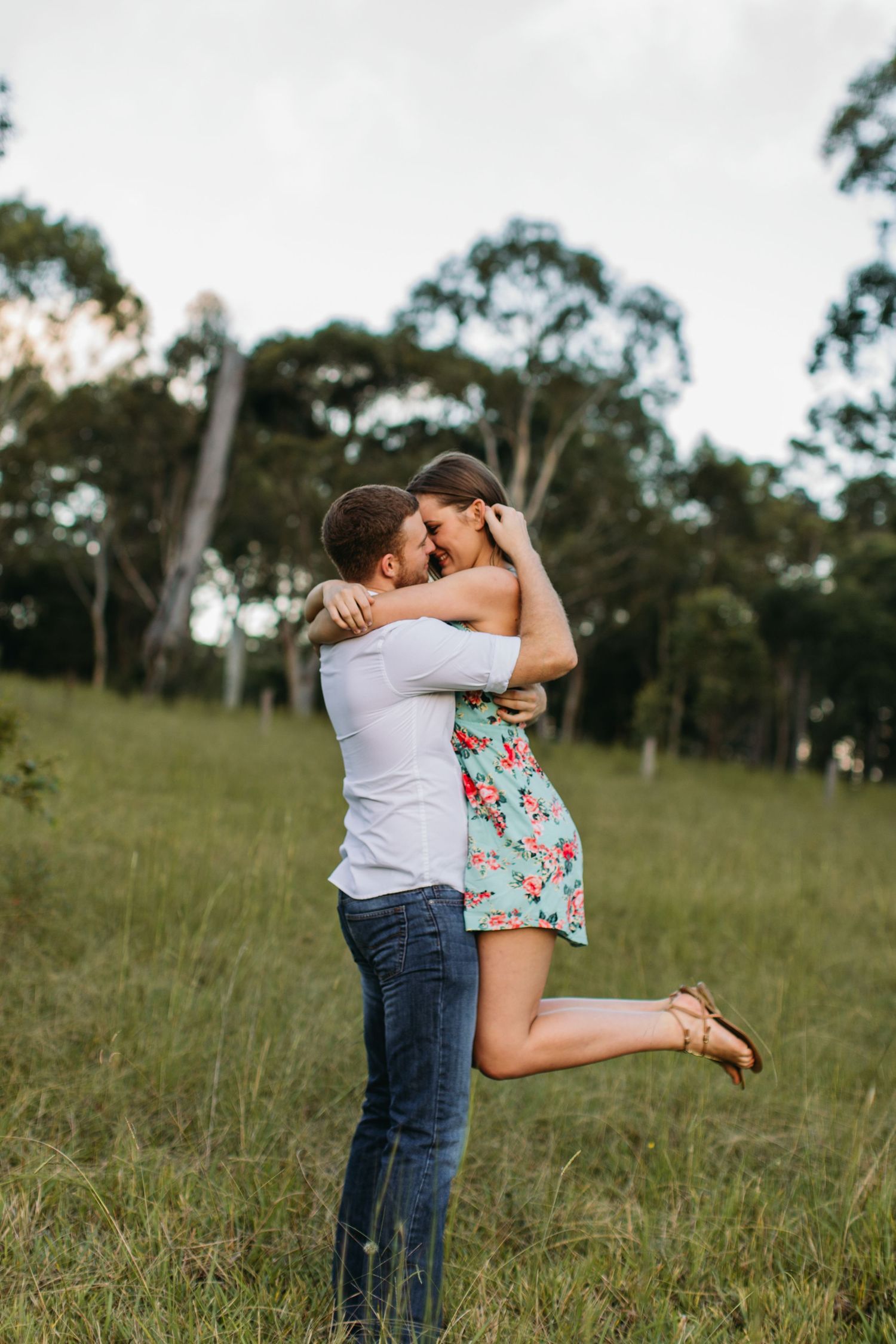 Couple embracing in a green field, man lifting woman in floral dress, trees in background.