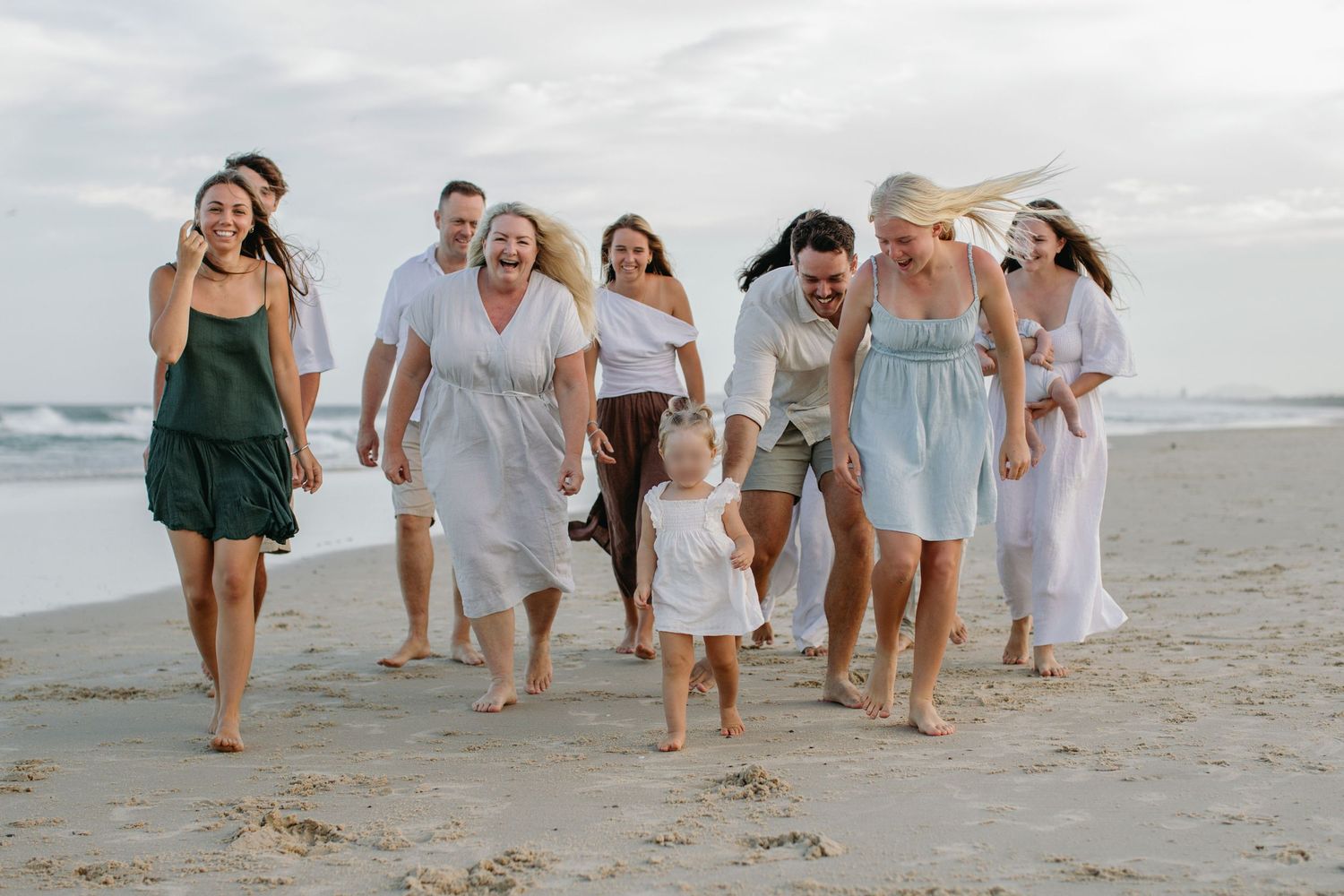 Large family group walking barefoot on a sandy beach, smiling and laughing together on a cloudy day.
