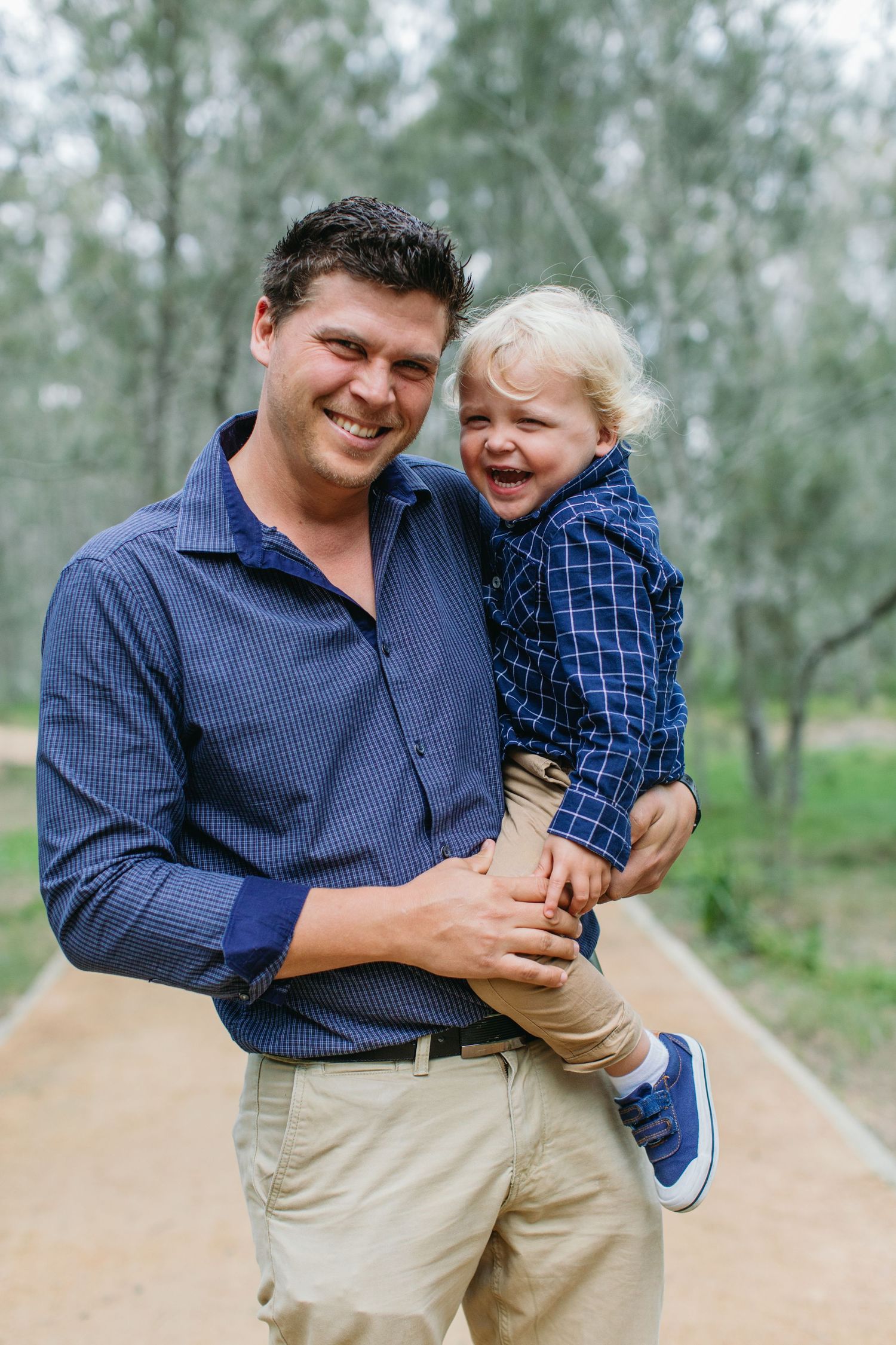 Smiling father holding laughing toddler boy outdoors on a dirt path surrounded by trees.