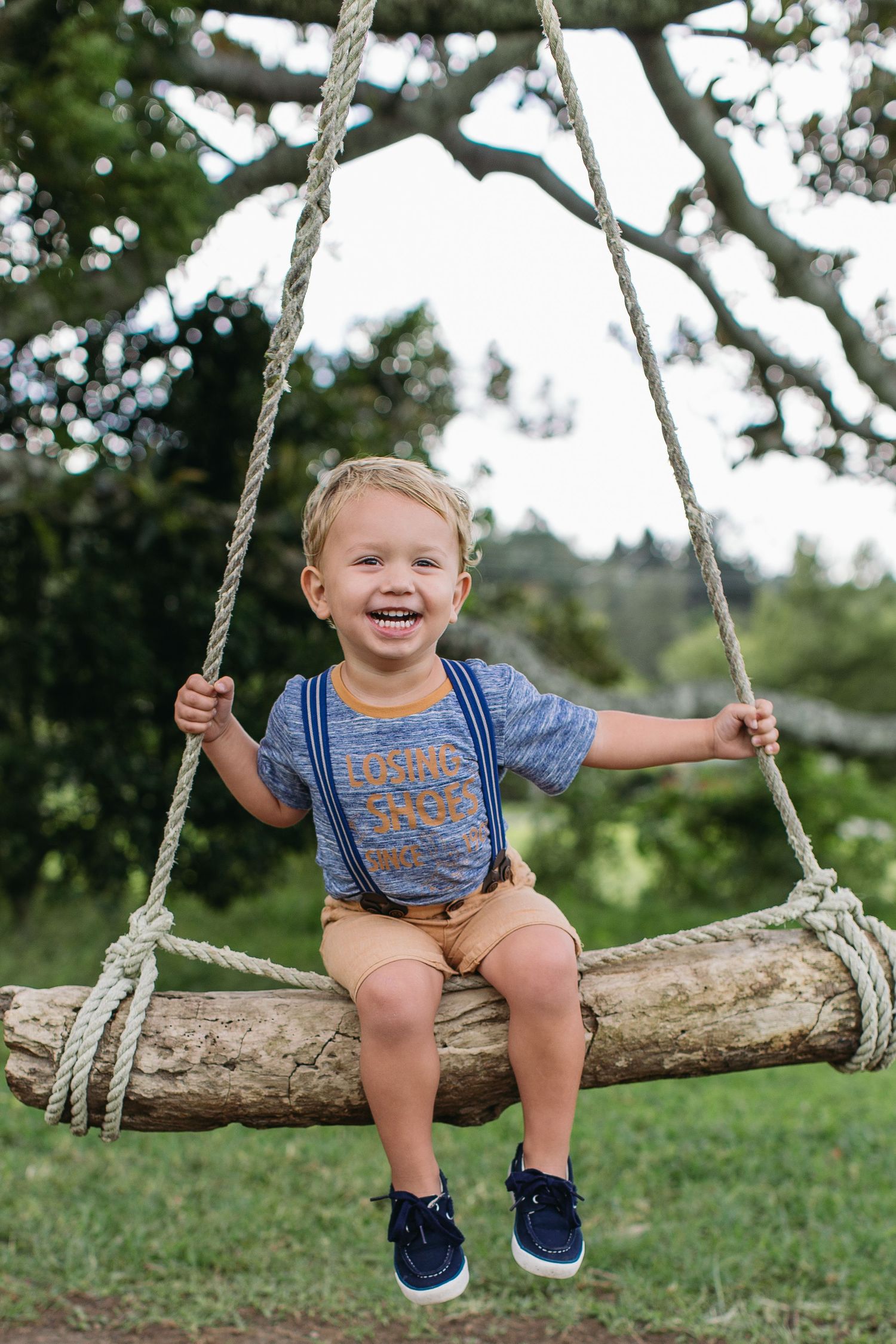Happy toddler boy sitting on a rustic rope tree swing outdoors, wearing suspenders and a blue shirt.