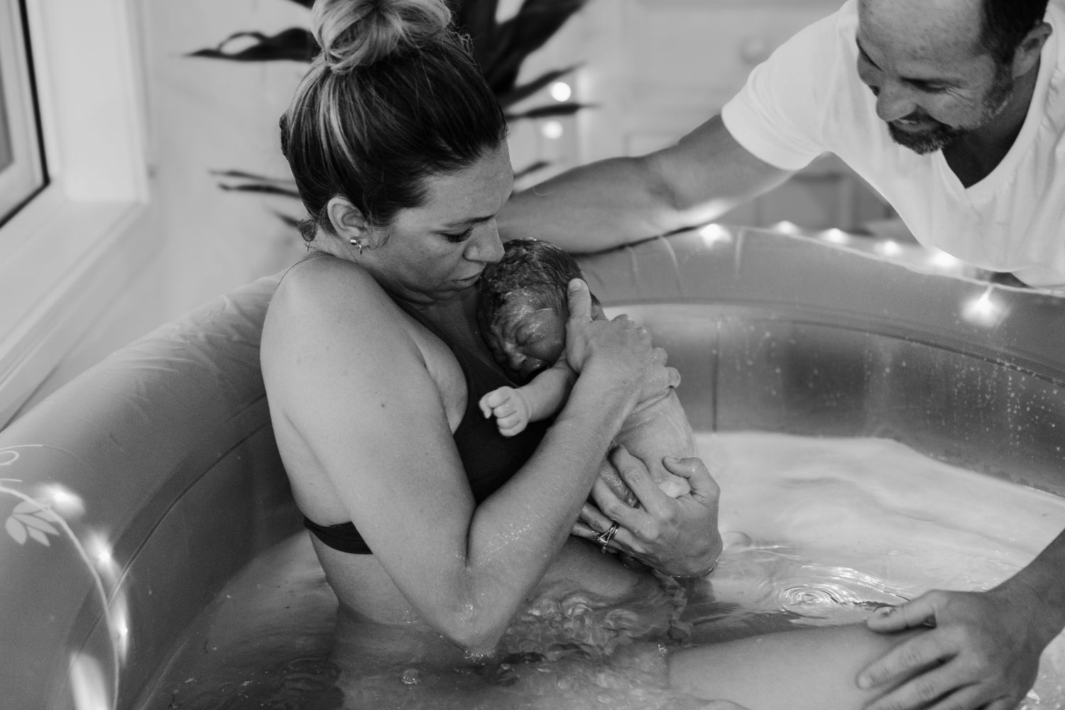 Black and white photo of mother lifting newborn from water birth pool with partner helping.