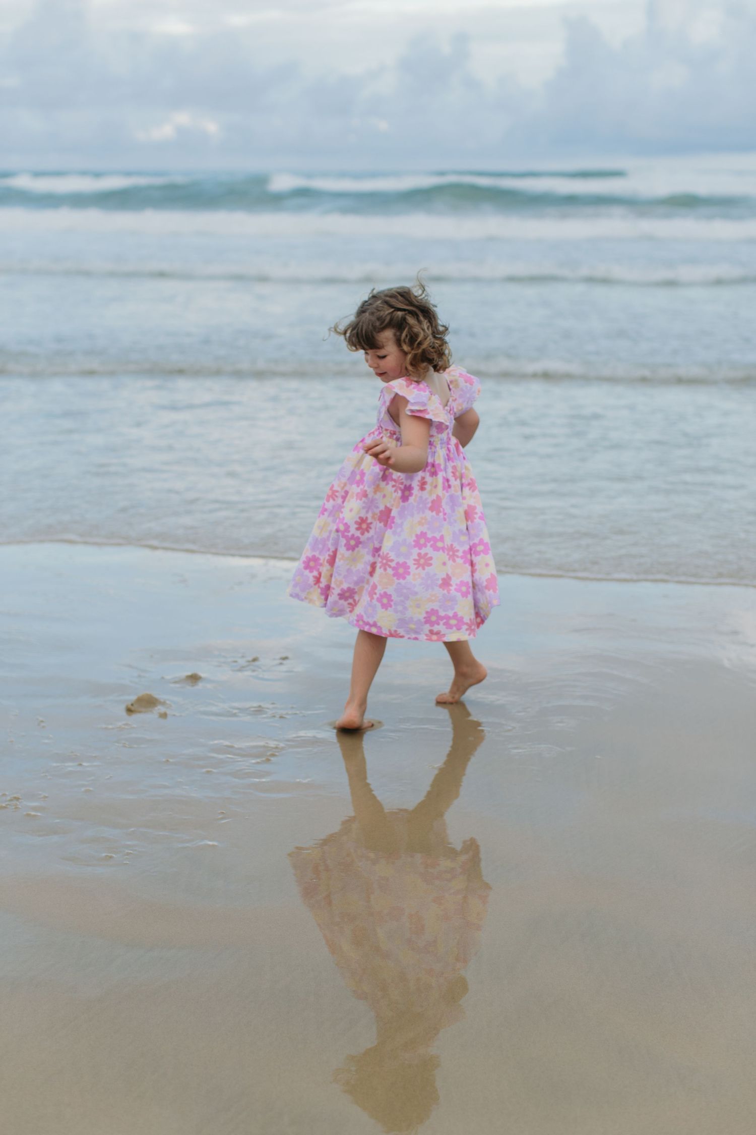 A young girl in a pink floral dress walks barefoot on wet sand at the beach, her reflection visible below.