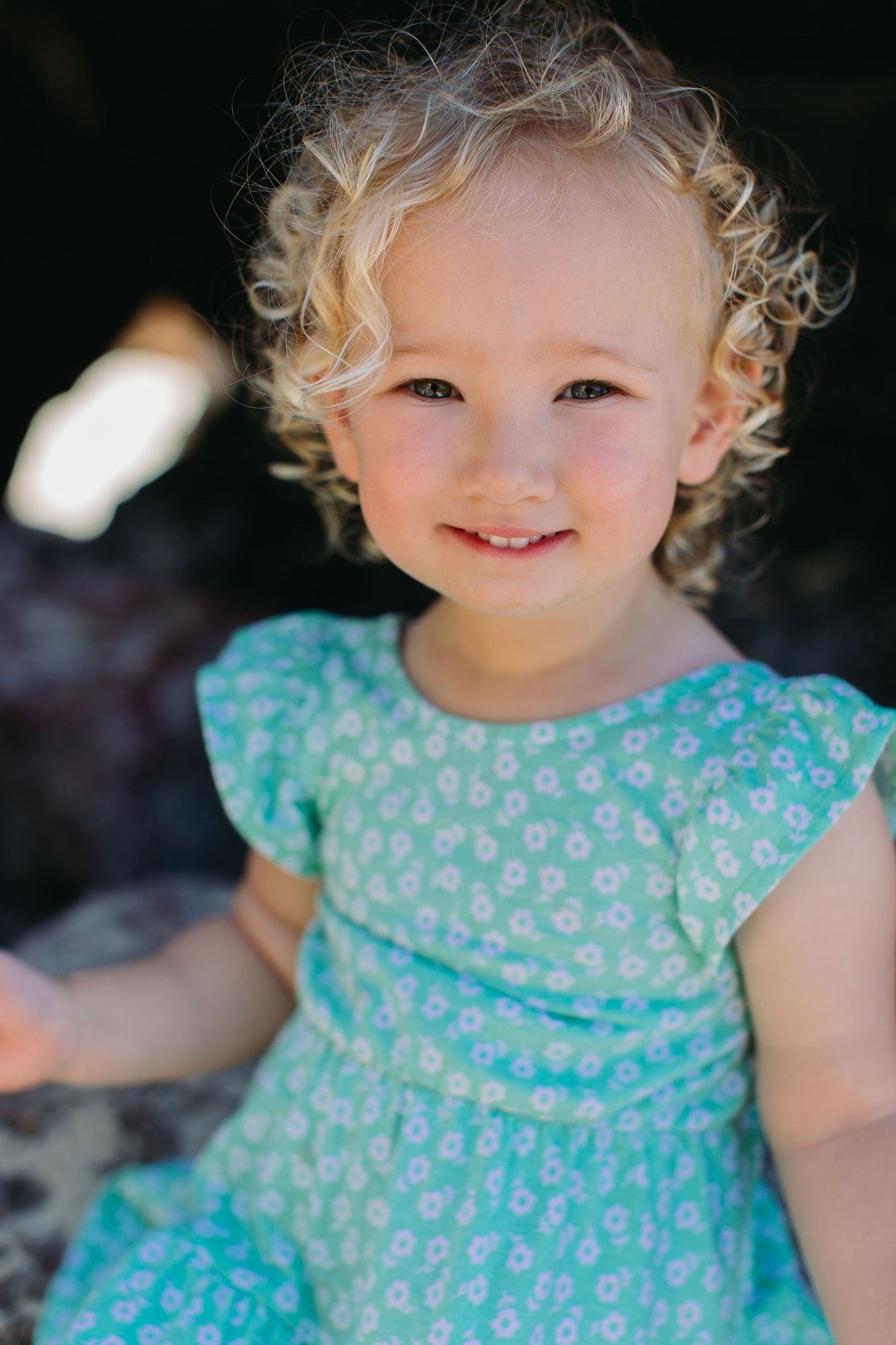 Adorable curly-haired blonde toddler girl smiling brightly in a teal floral dress outdoors in natural sunlight.