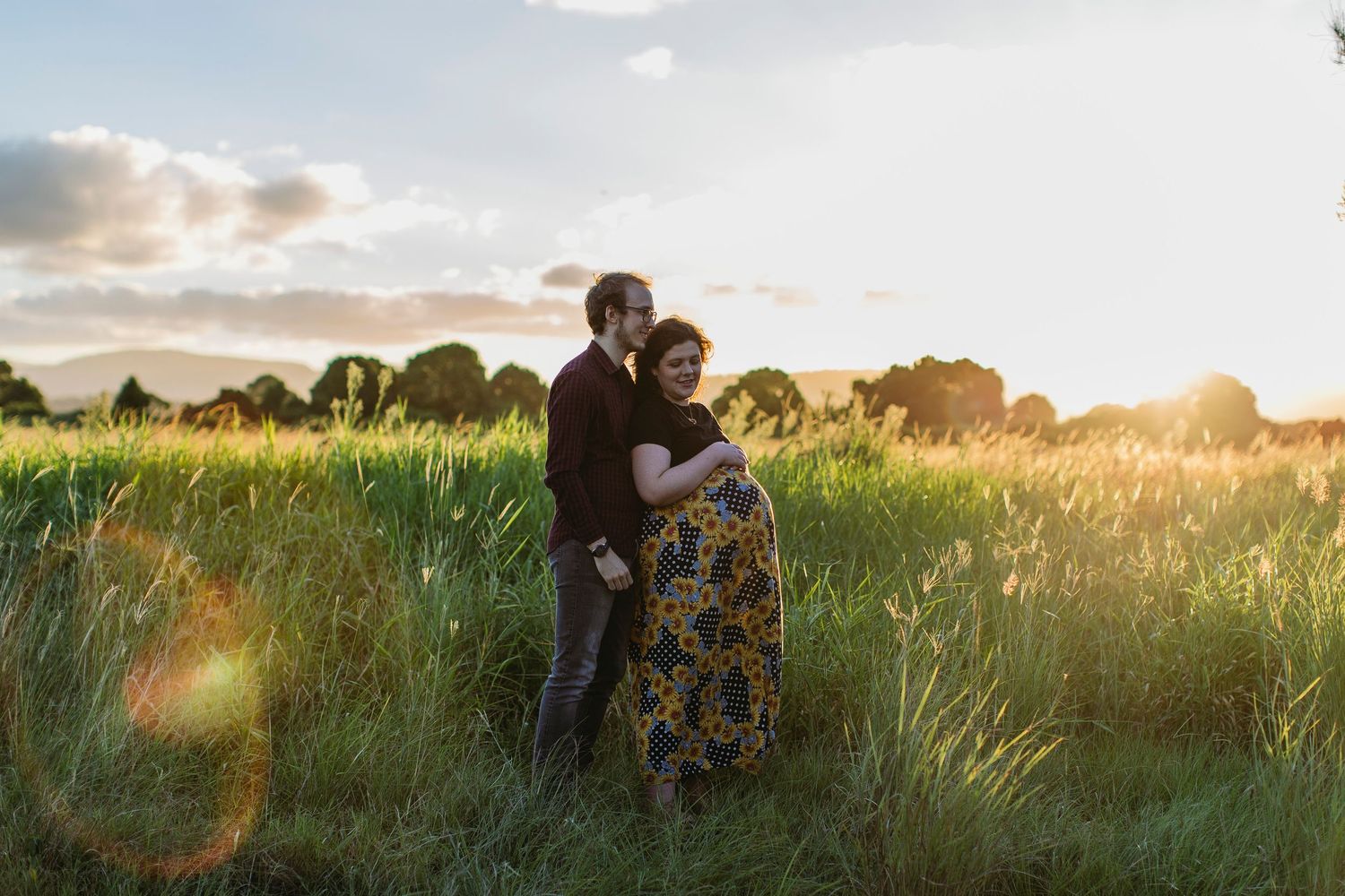 Couple embracing in golden sunset field, pregnant woman in floral dress, romantic maternity photo session outdoors.