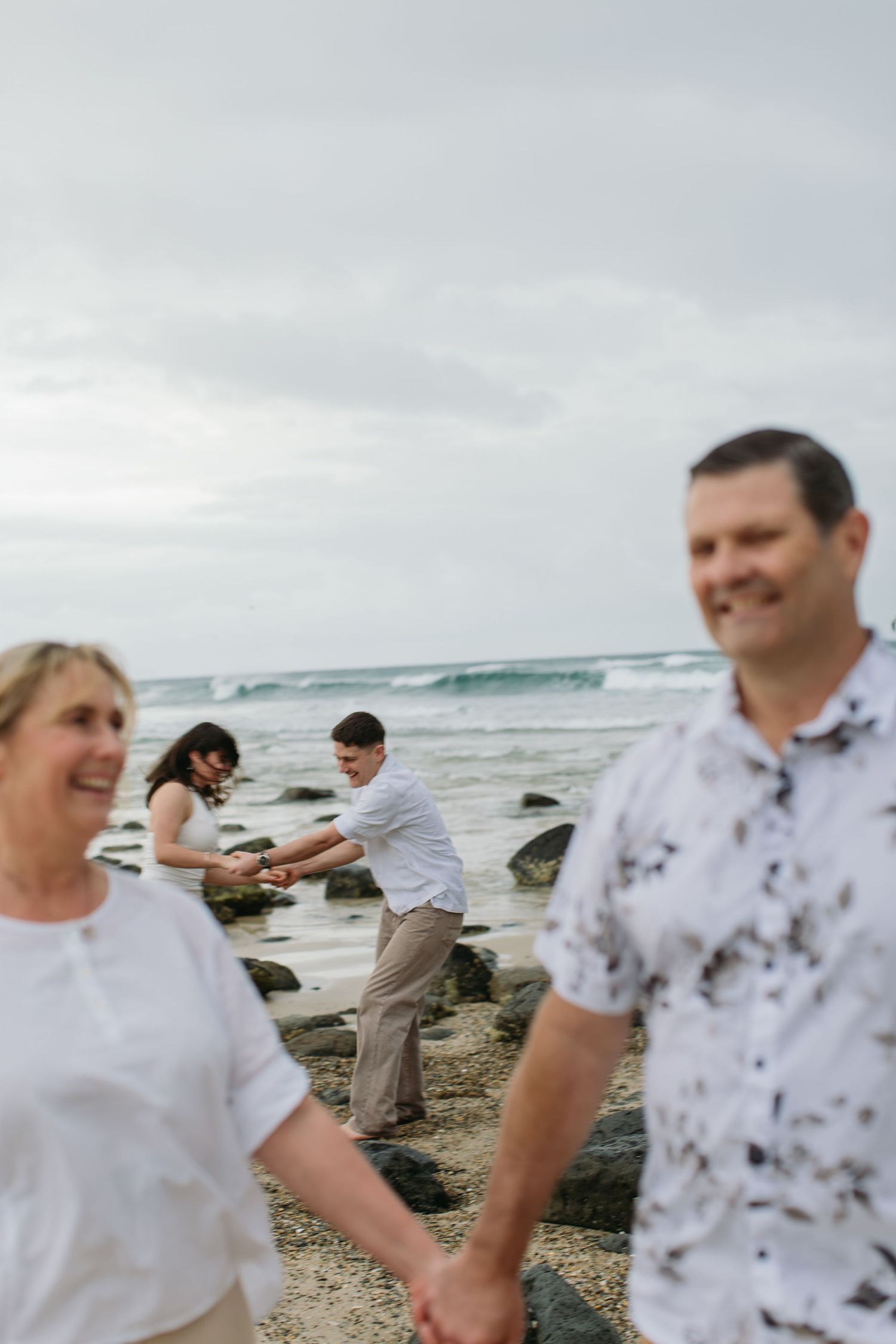 Two couples enjoy a romantic beach day, holding hands and playing near rocky shores with ocean waves in the background.