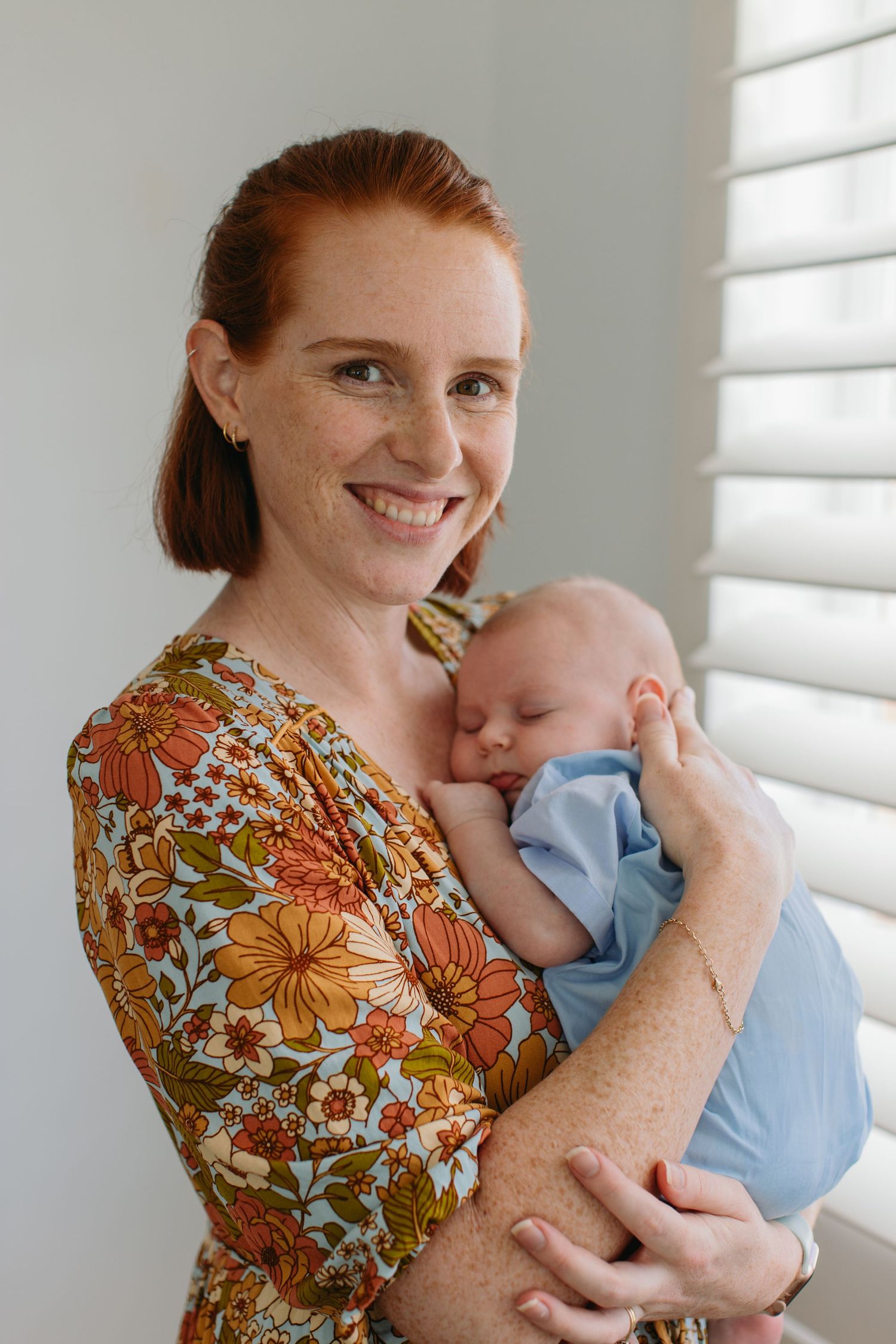 Smiling redhead mother in floral dress tenderly holds sleeping newborn baby in blue onesie near window.