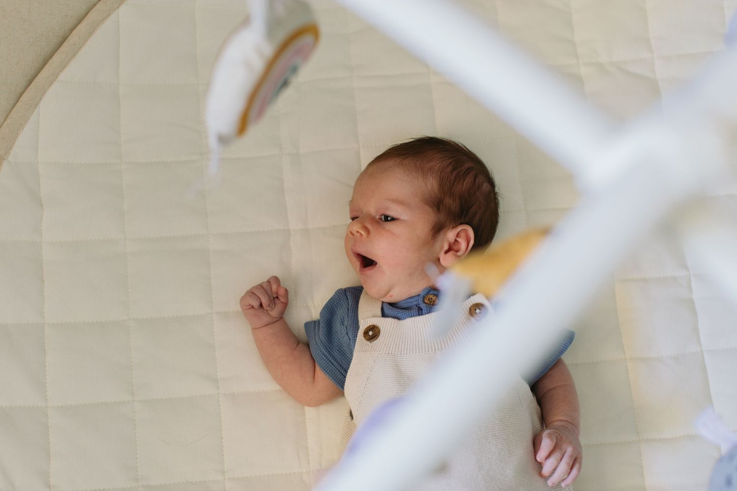 Crying newborn baby lying on back in white crib with mobile overhead, wearing denim romper.