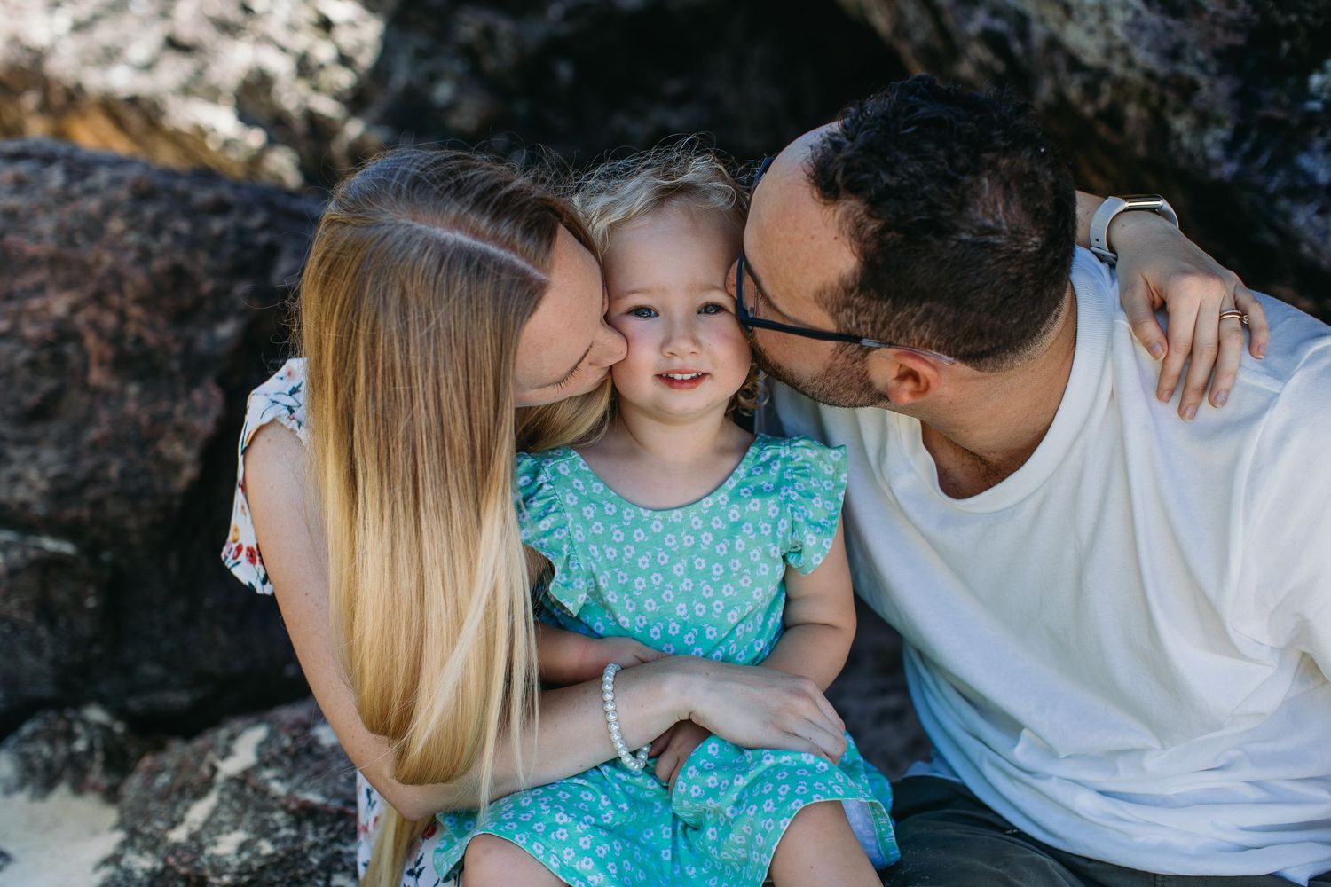 Loving parents kissing their smiling toddler daughter on cheeks at rocky beach setting.
