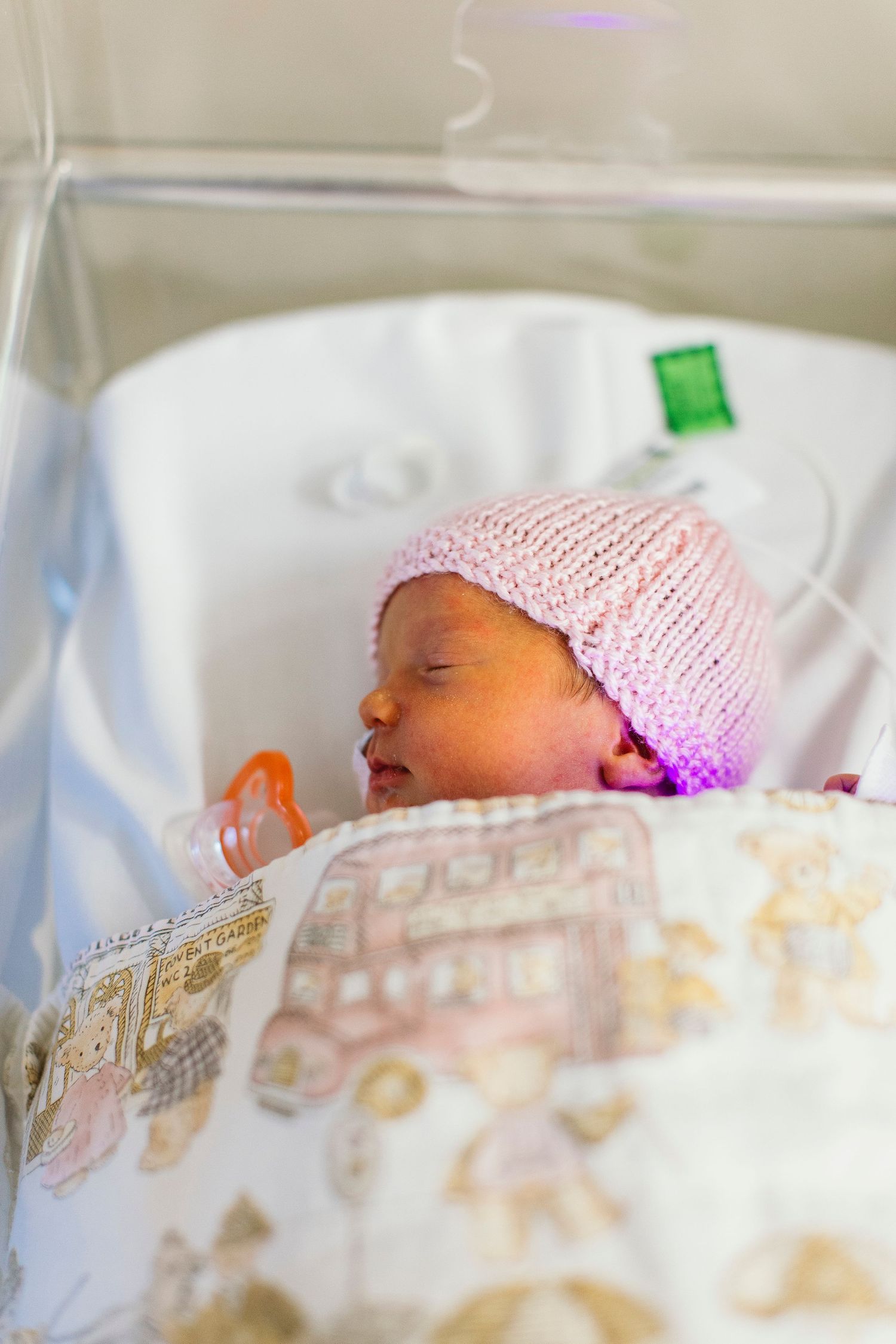 Newborn baby sleeping in hospital bassinet wearing pink knit hat with pacifier, wrapped in decorative blanket.
