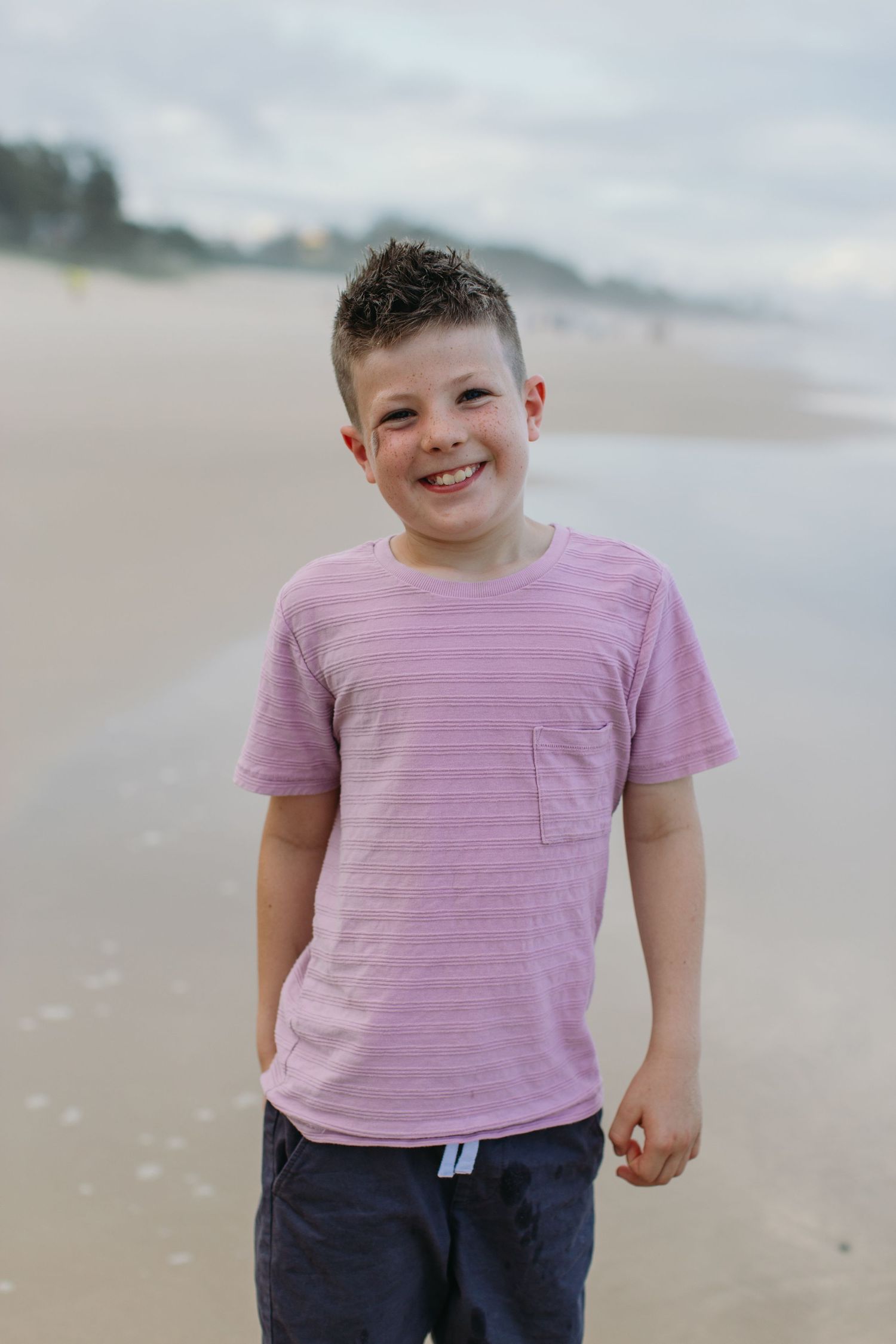 A smiling boy in a pink striped t-shirt stands on a sandy beach under a cloudy sky.