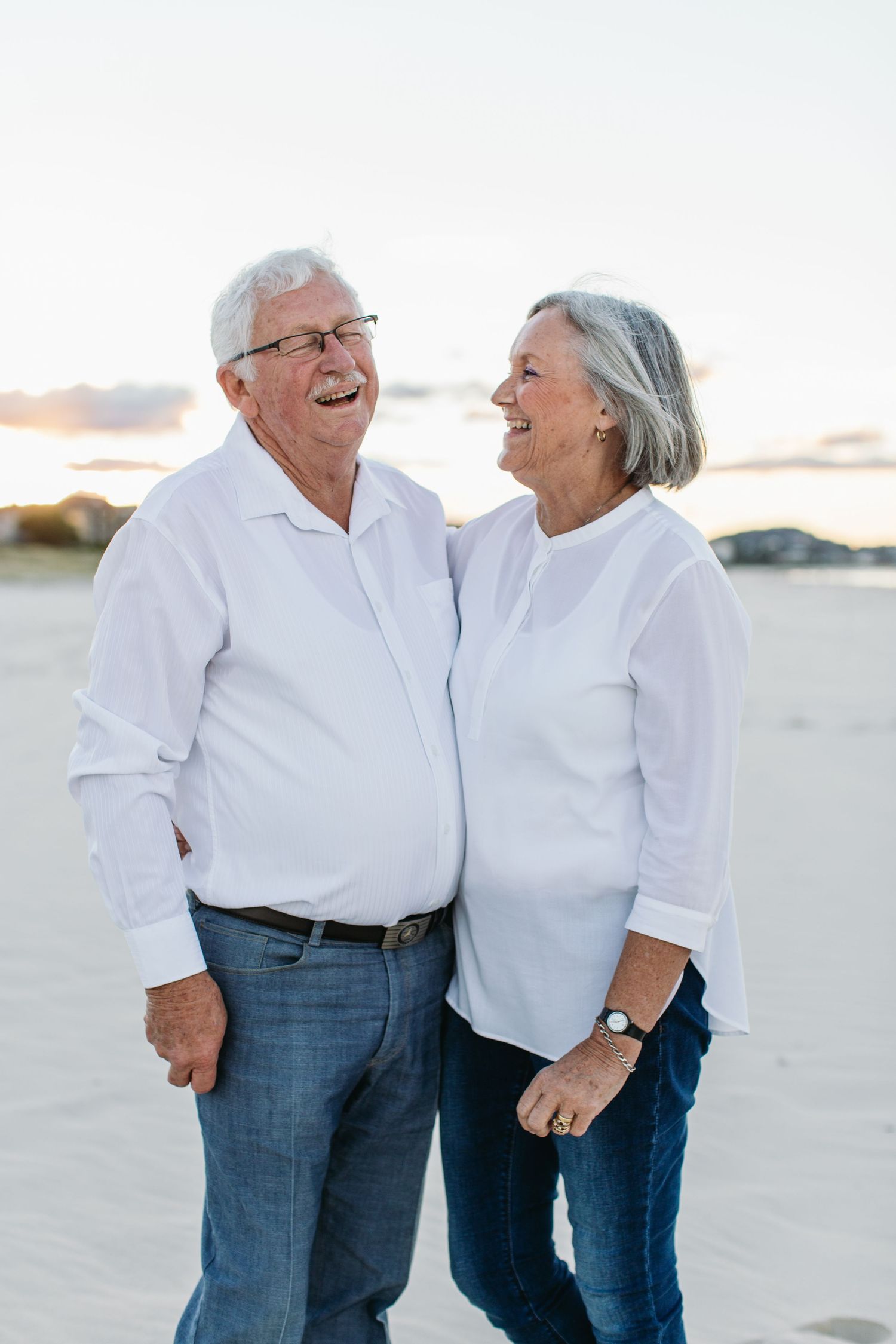 Happy elderly couple in white shirts laughing together on a sandy beach at sunset.