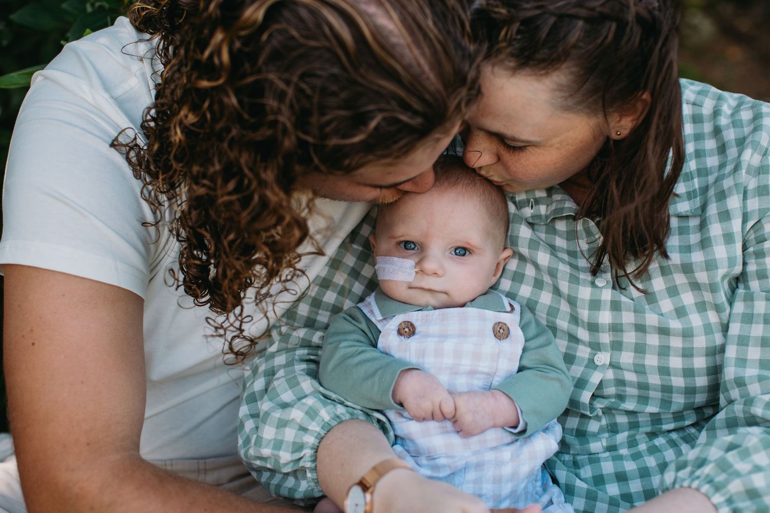Two women kissing a cute baby dressed in blue gingham outfit, sharing a tender family moment outdoors.