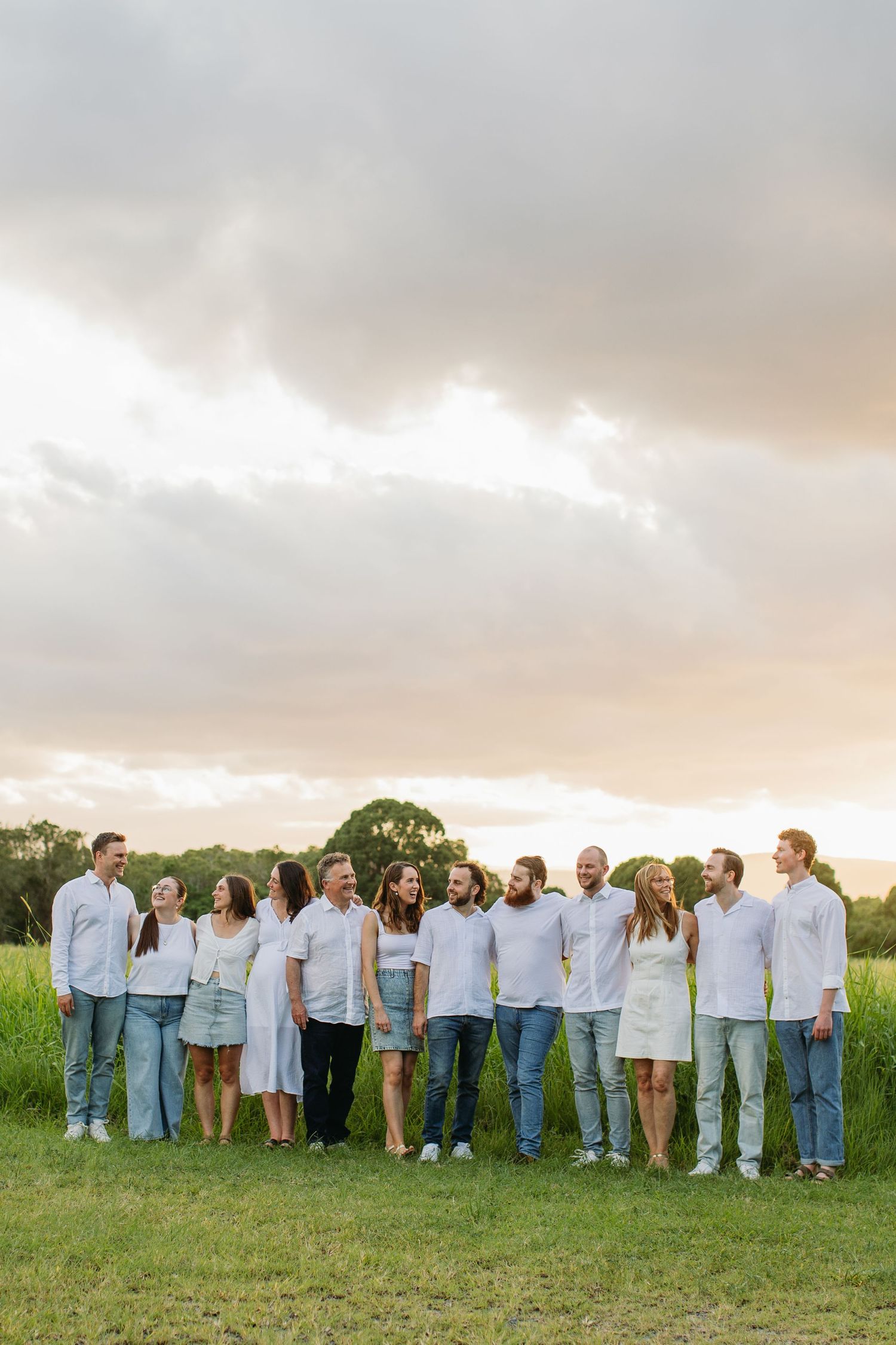 Large family group posing together in white shirts and jeans in a green field at golden hour sunset.