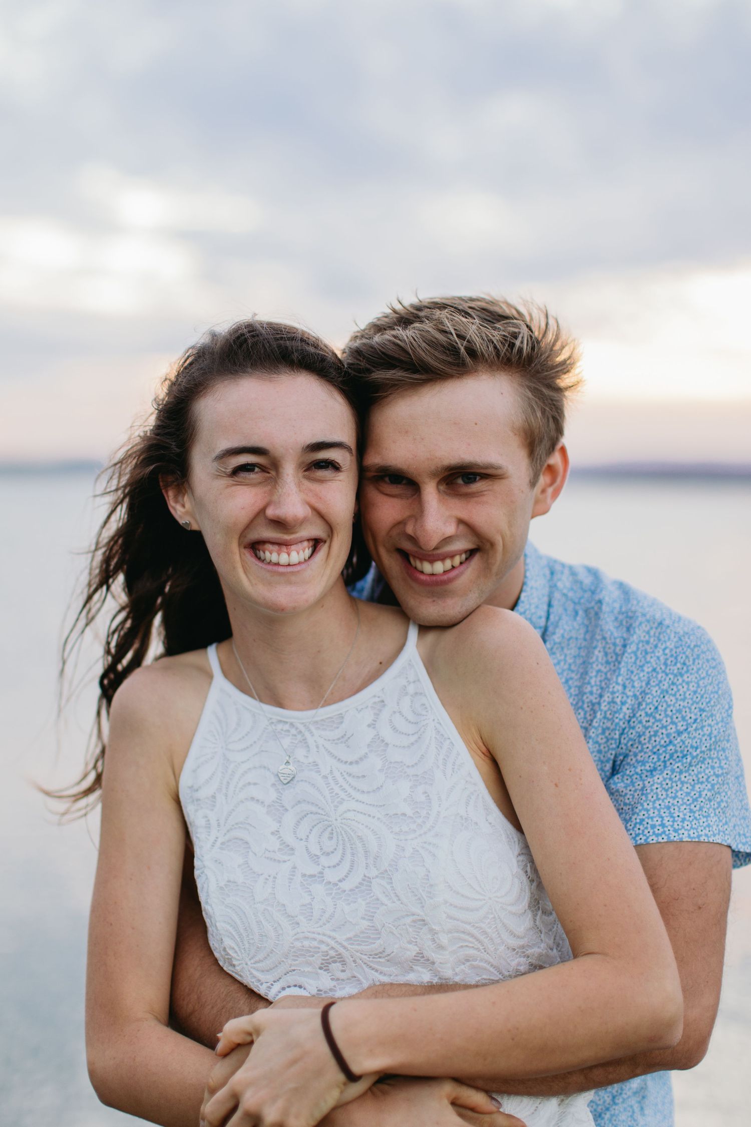Happy young couple embracing on a beach at sunset, smiling joyfully with ocean and cloudy sky behind them.