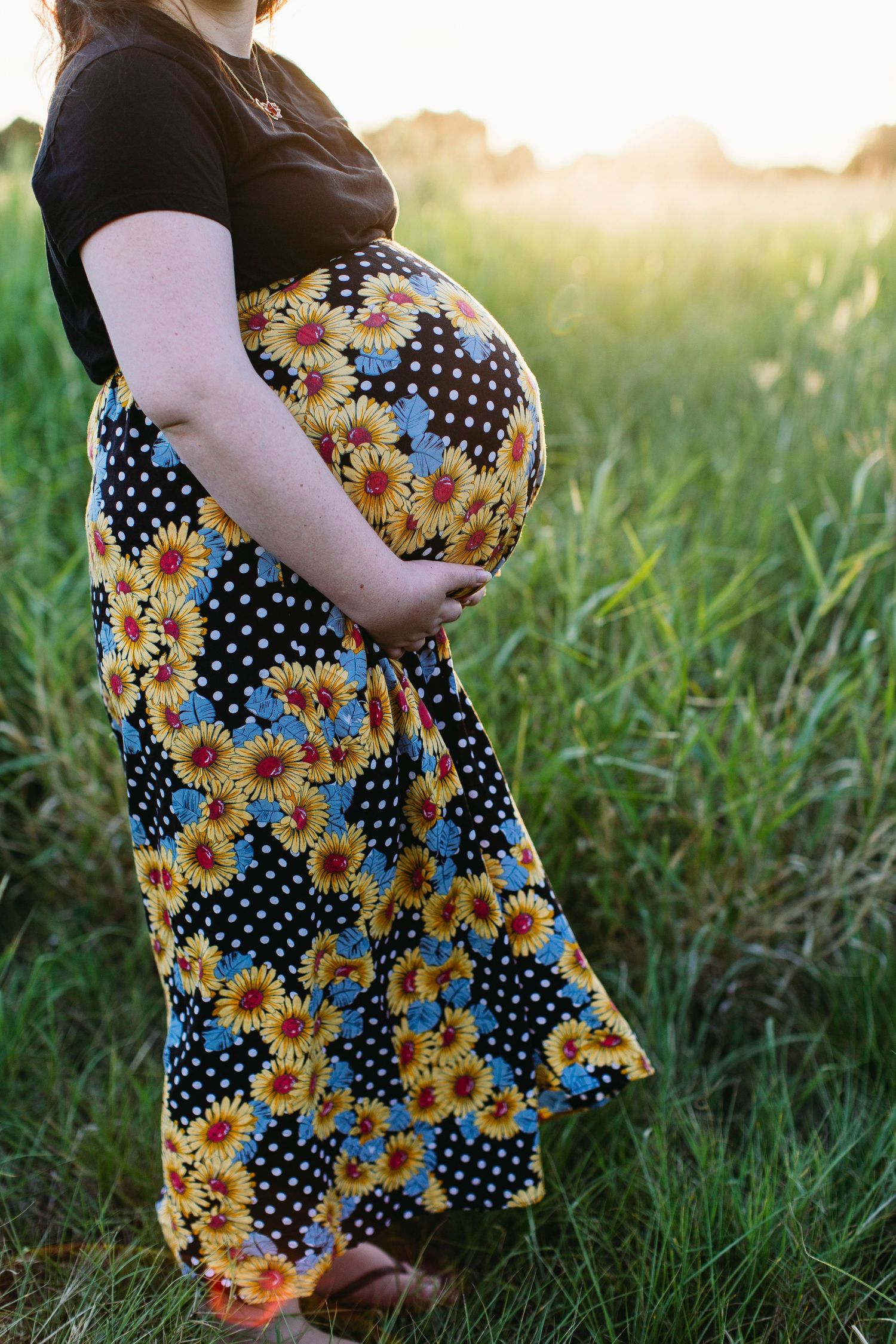 Pregnant woman in floral skirt holding baby bump in sunlit green field at golden hour.