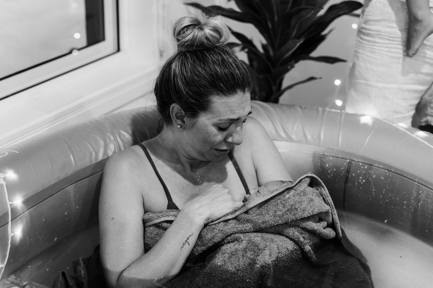 Black and white photo of a mother cradling her newborn in a home water birth pool with fairy lights.