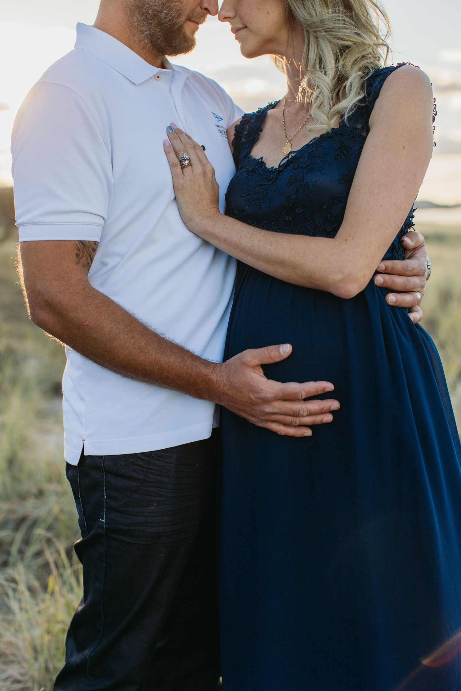 A couple shares a tender moment outdoors, with the man gently holding his pregnant partner's belly in a navy maternity dress.