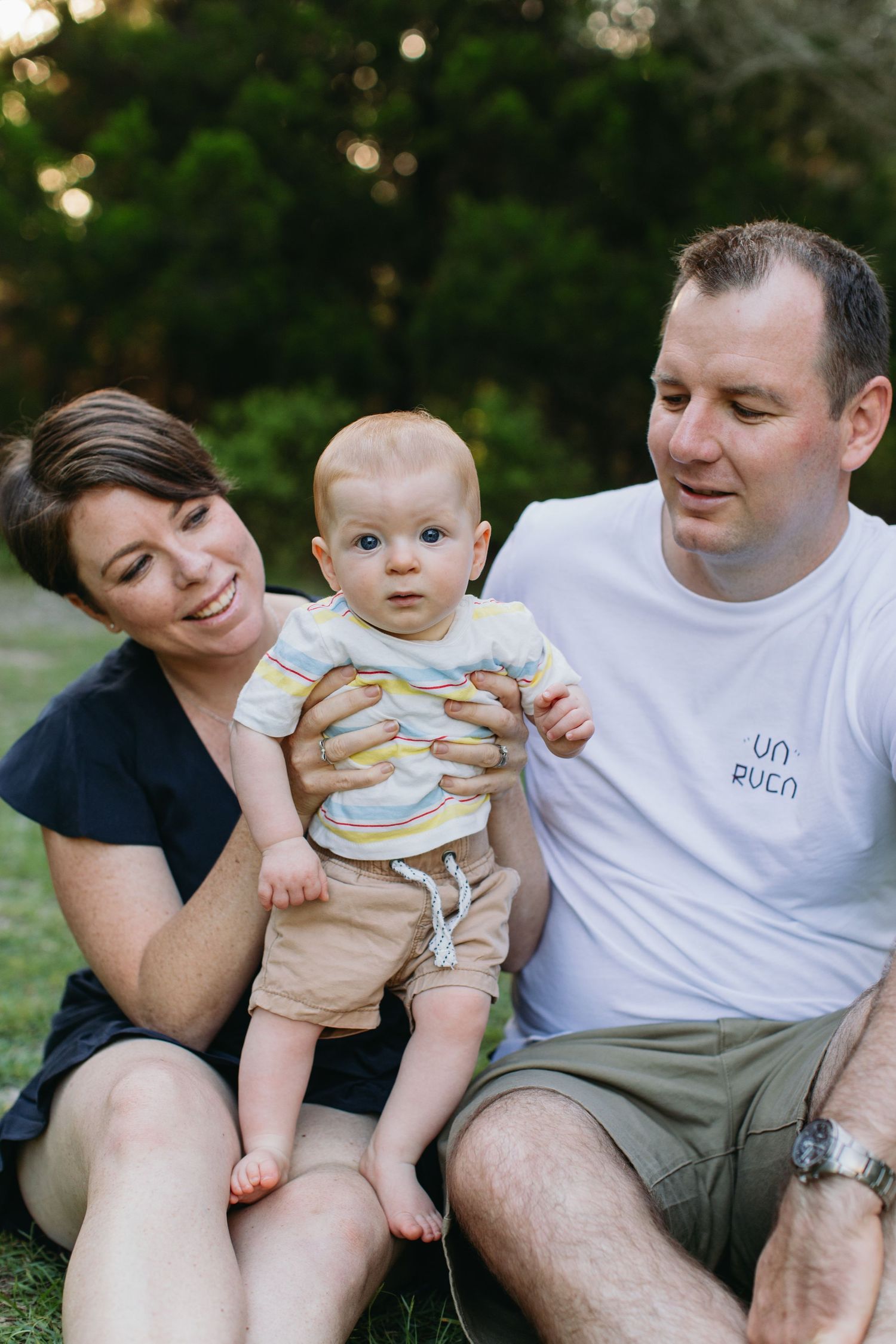 Happy family with parents sitting on grass outdoors holding their curious baby boy in colorful striped outfit.
