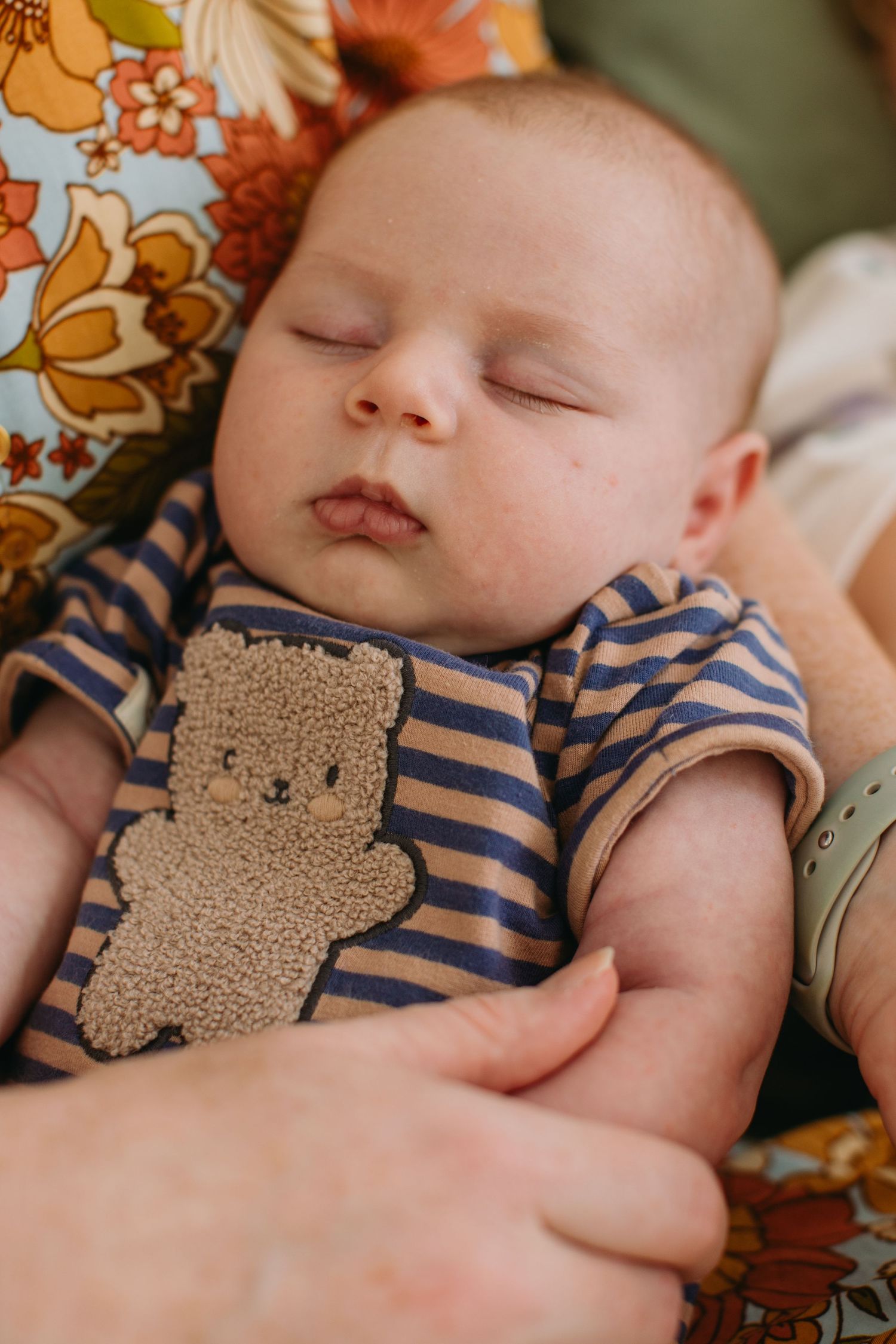Sleeping newborn baby in striped bear onesie cradled in adult arms on floral background.