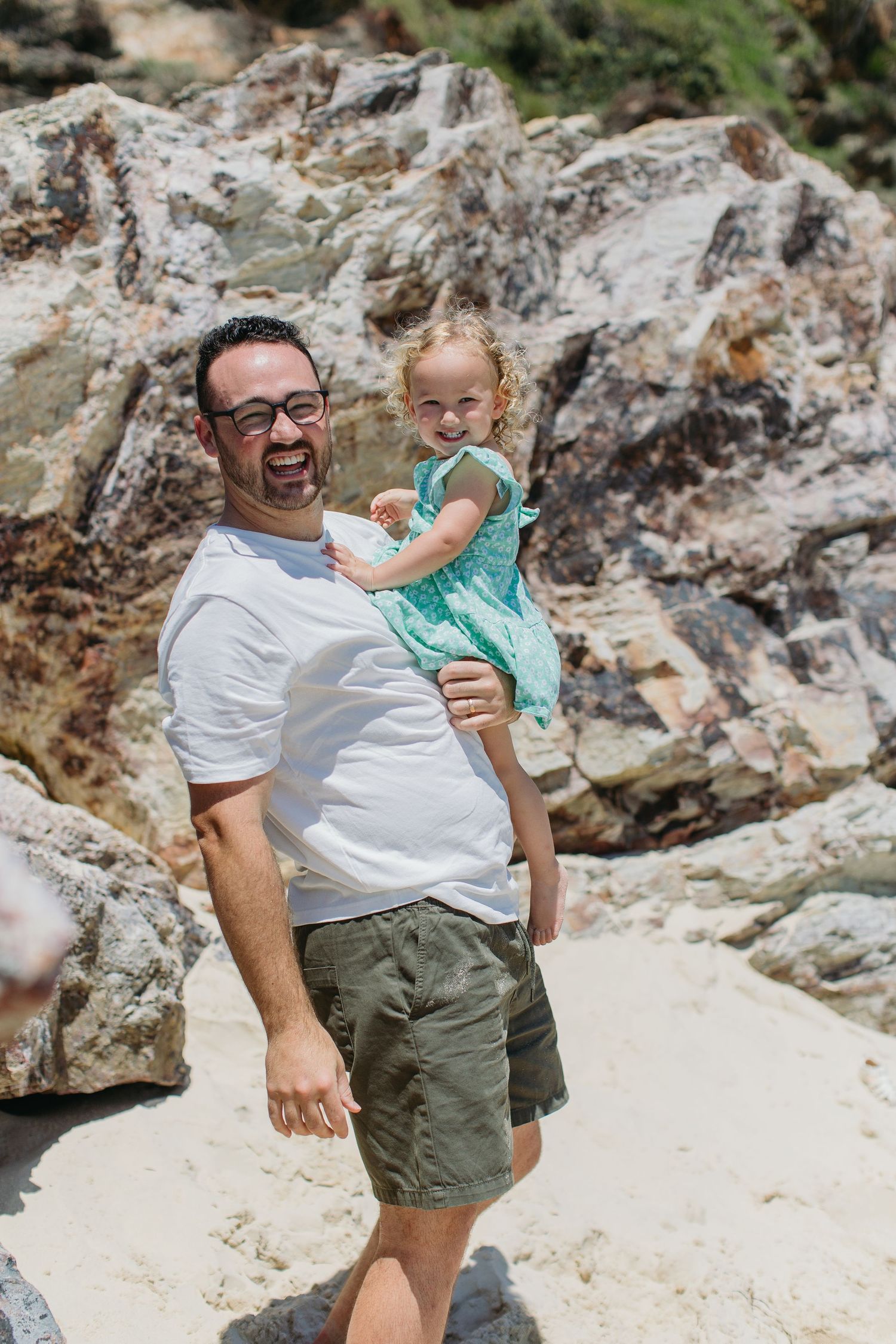 Joyful dad in white tee lifting giggling toddler in teal dress on sunny rocky beach.