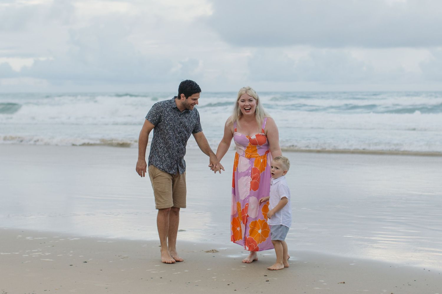 Family of three walking hand-in-hand on a beach, waves in background, woman in colorful dress, man in casual wear.