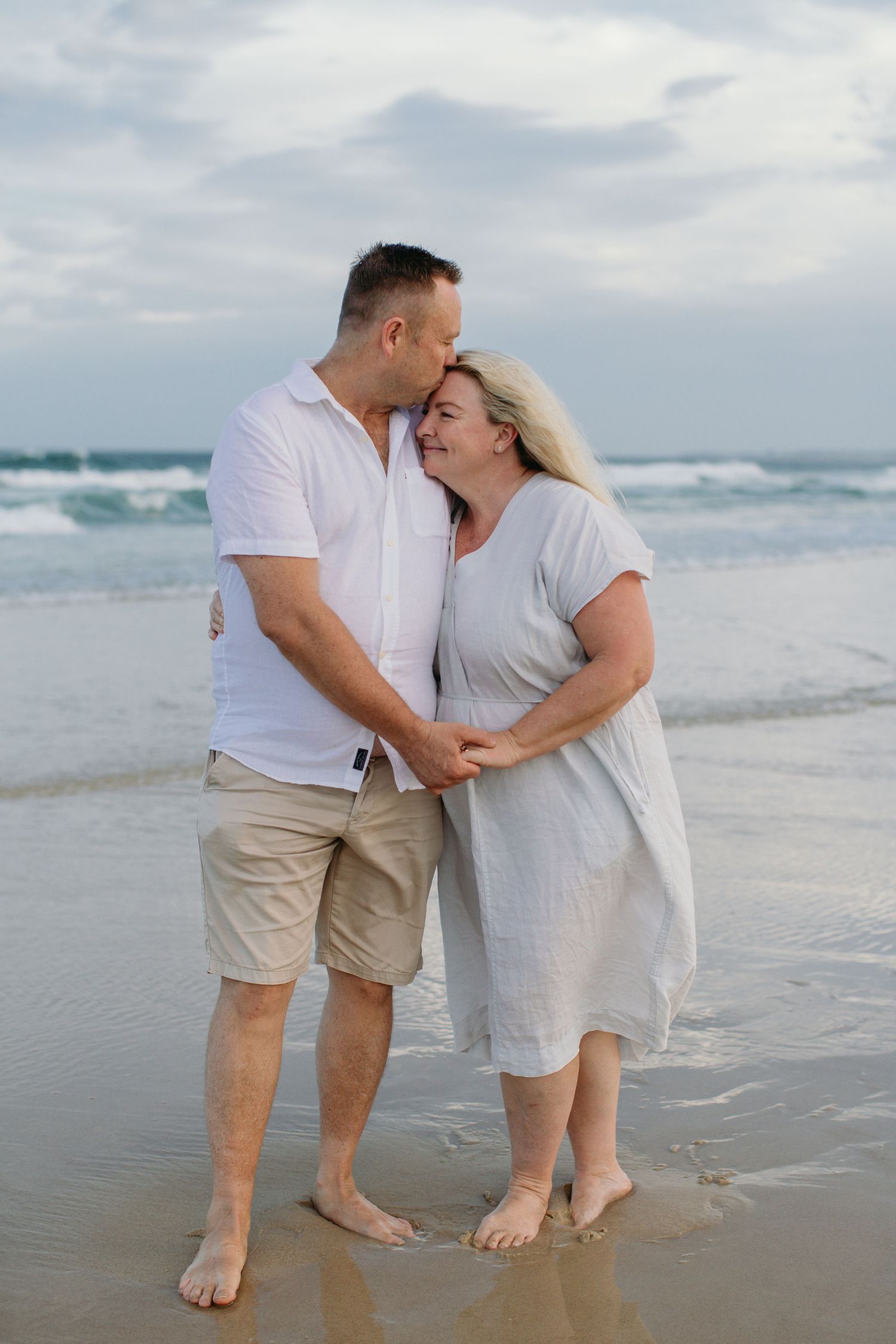 Couple holding hands and sharing a kiss on a sandy beach with ocean waves in the background.