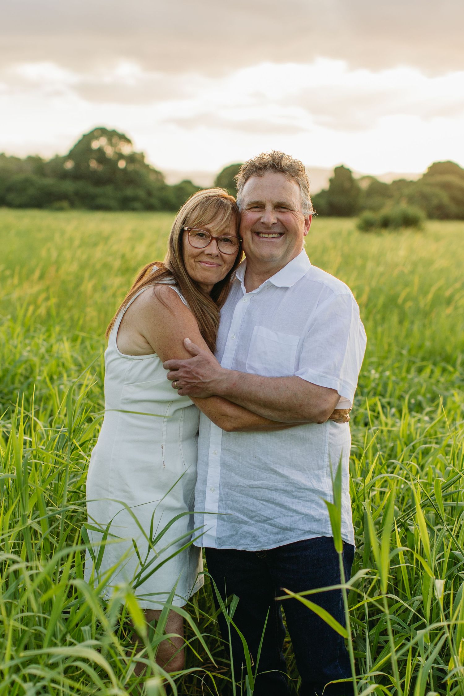A smiling couple embraces in a lush green field at golden hour, woman in white dress, man in white shirt.