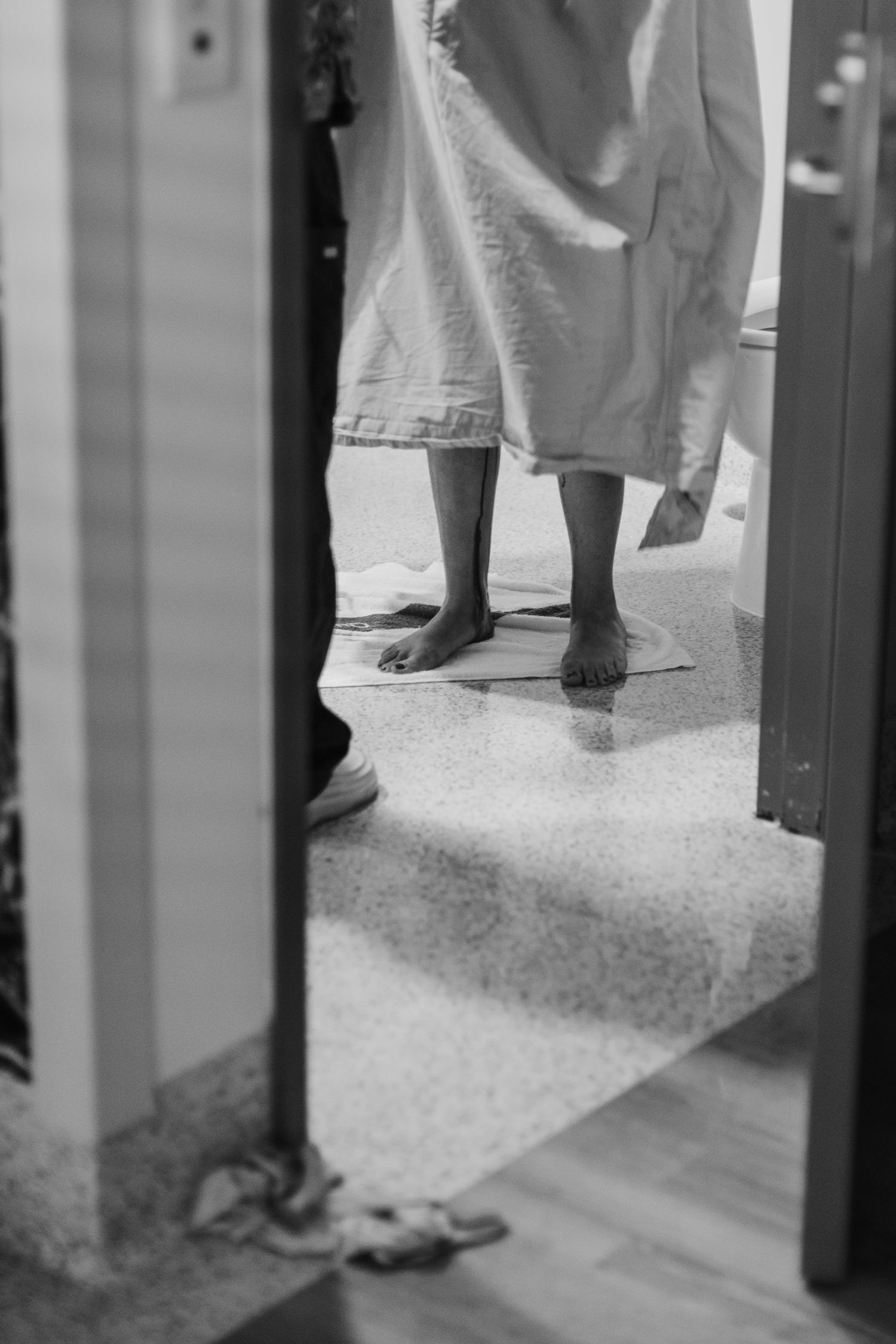 Black and white photo of a patient in hospital gown standing barefoot on tiled floor, viewed through doorway.