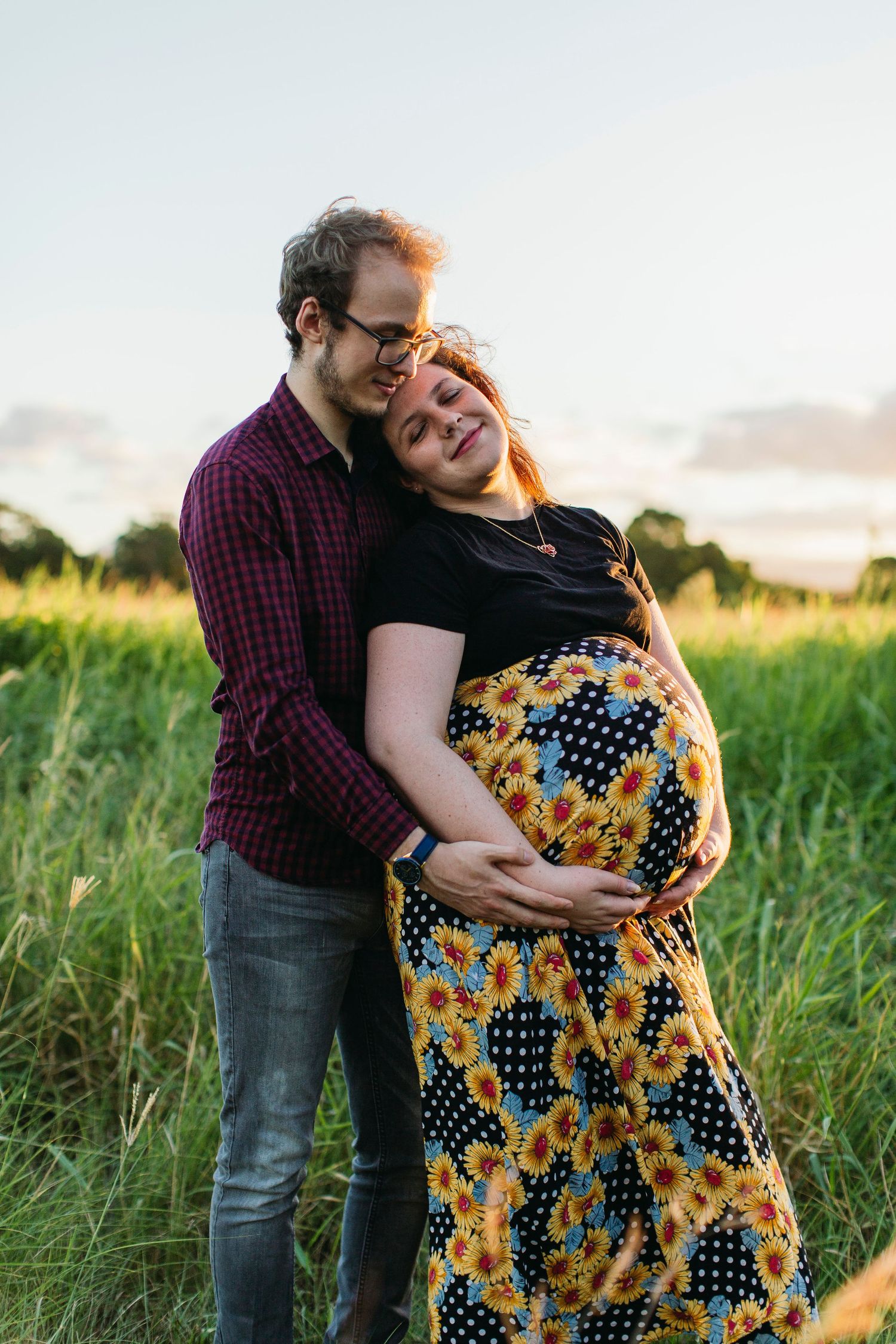 A couple embraces in a green field at sunset, the woman in a sunflower dress cradling her pregnant belly.