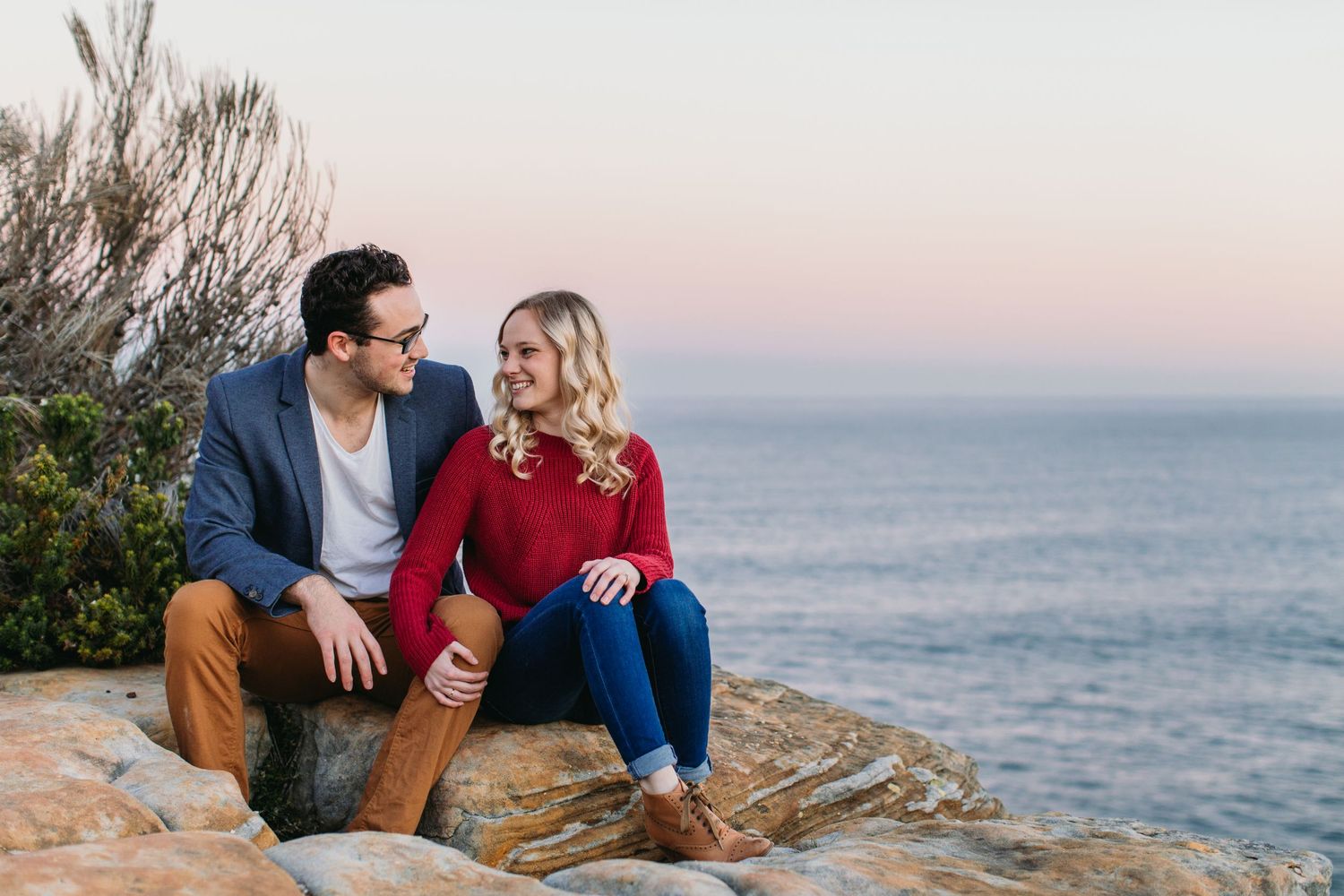 Couple sitting on coastal rocks at sunset, man in blue blazer and woman in red sweater sharing a romantic moment.