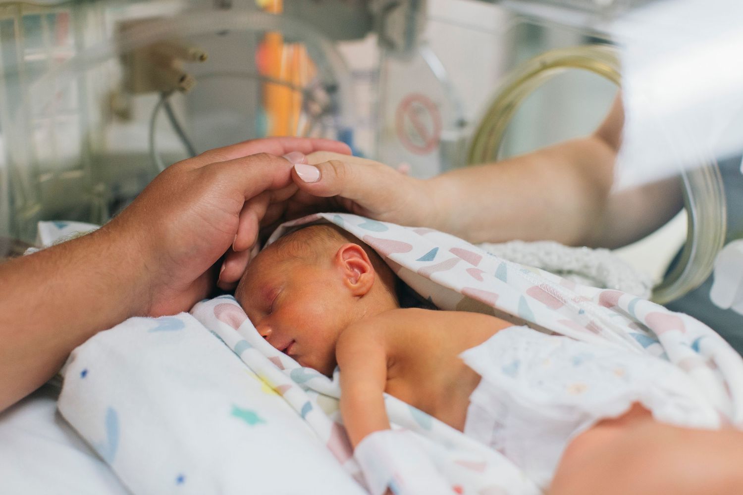 Newborn baby in NICU incubator with parent's gentle hand resting on head, wrapped in colorful blanket.