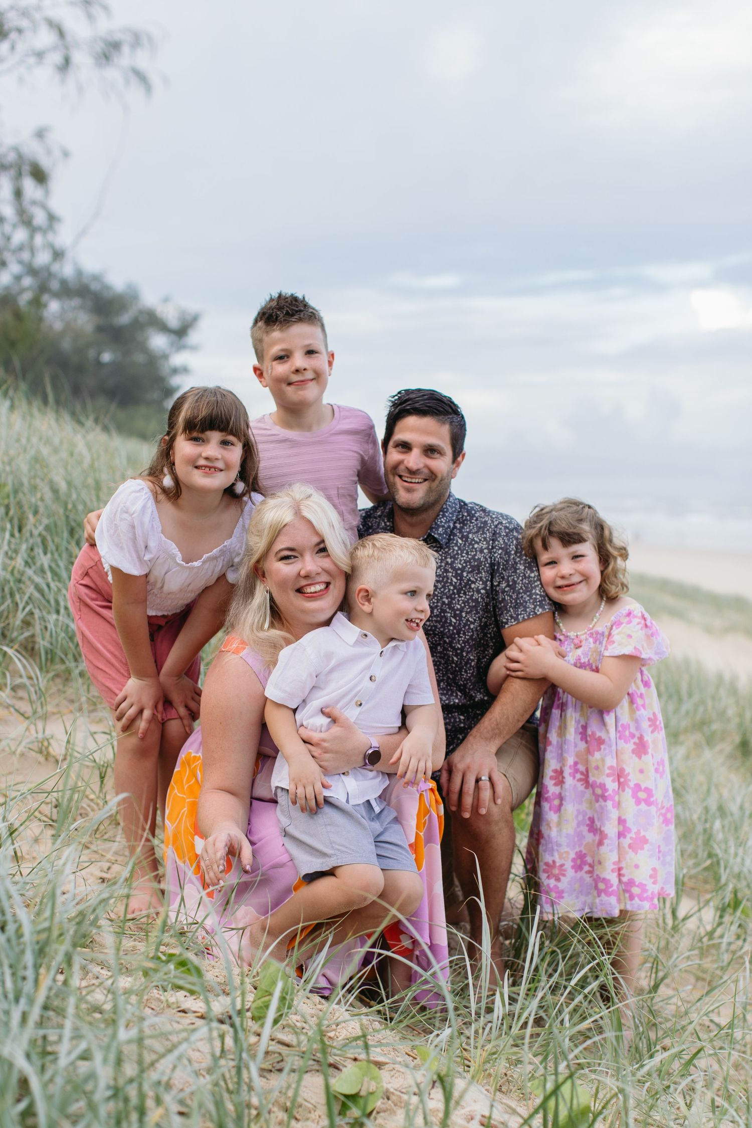 Family of six smiling together in beach sand dunes on cloudy day.