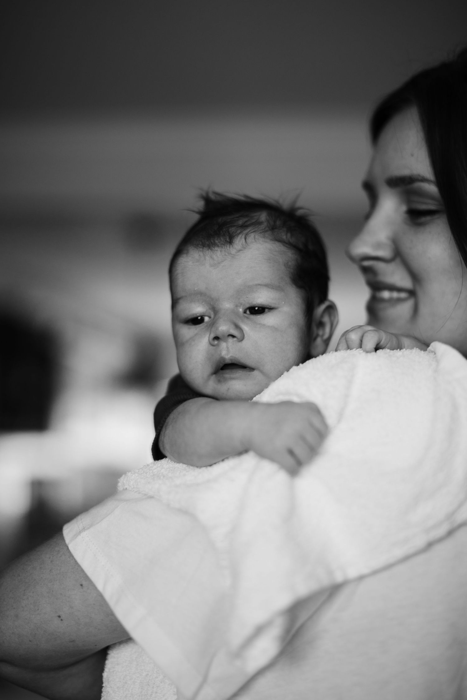 Mother smiling while holding newborn baby wrapped in white towel, black and white portrait.