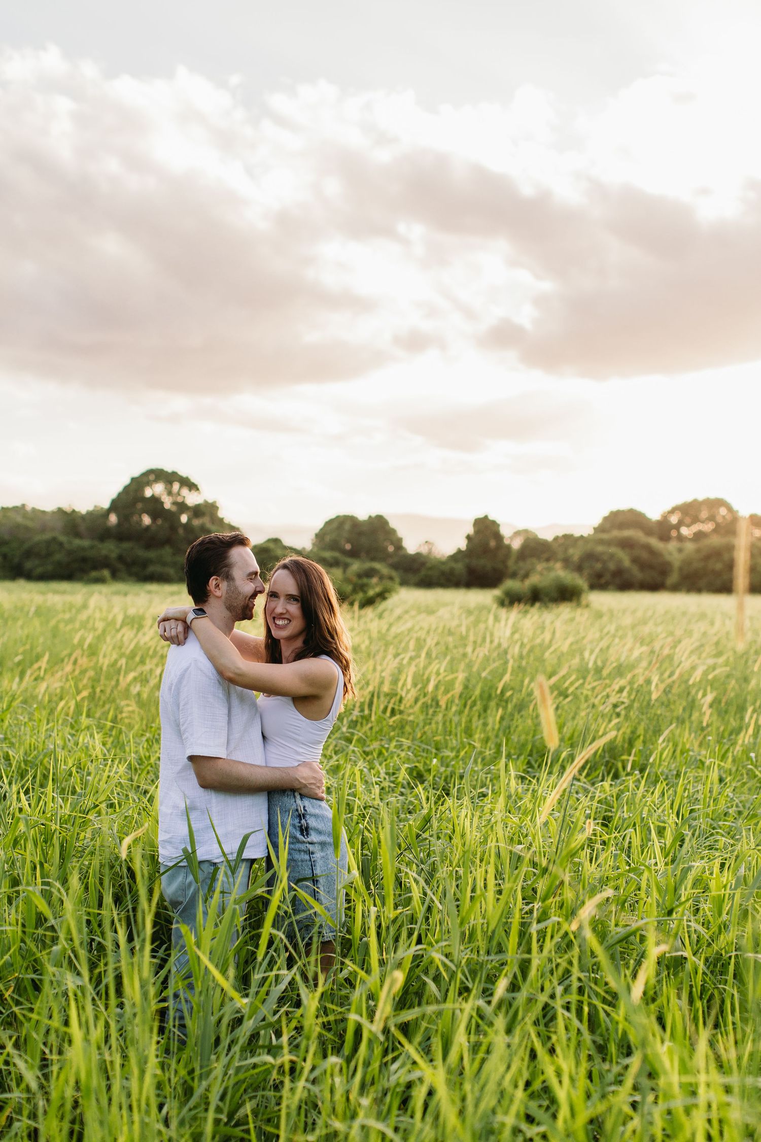Happy couple embracing and laughing in a lush green field at golden hour sunset.