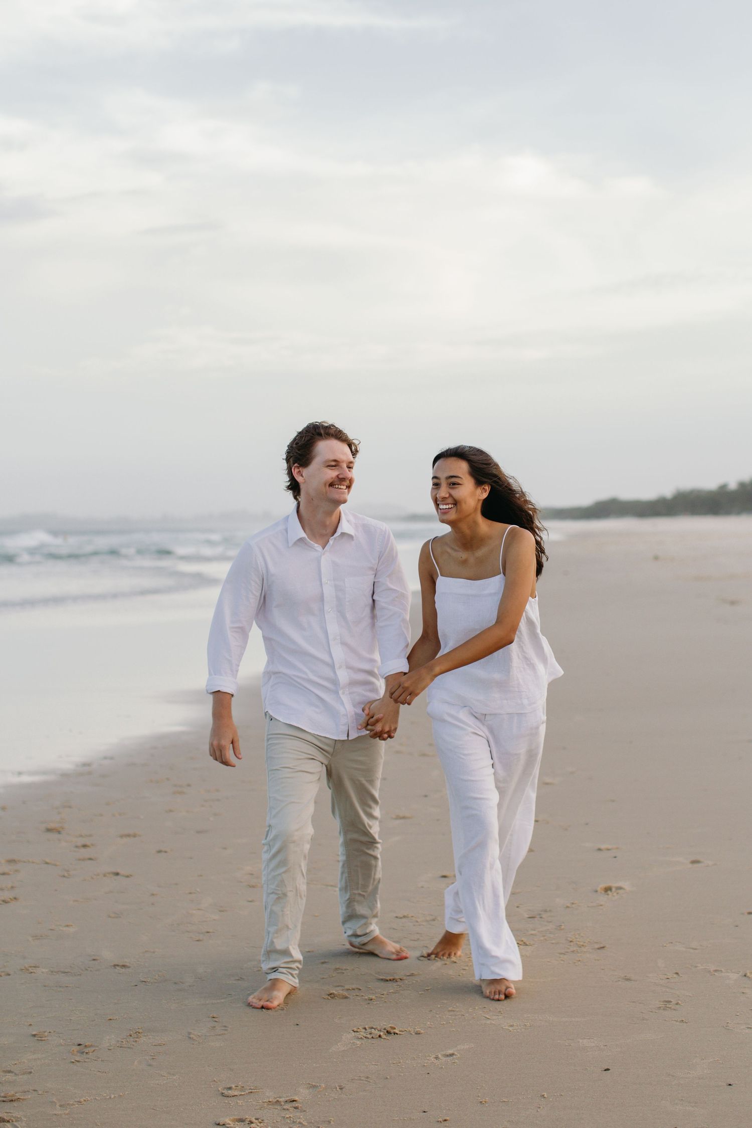 Happy couple in white outfits holding hands and walking barefoot on a sandy beach with ocean waves in the background.
