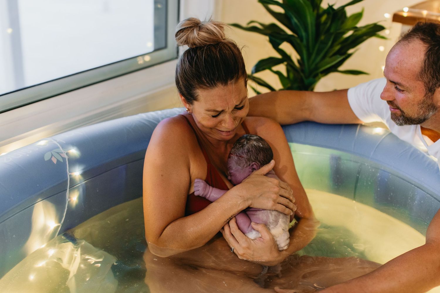 A mother holds her newborn baby in a water birth pool while a supportive partner assists beside her.