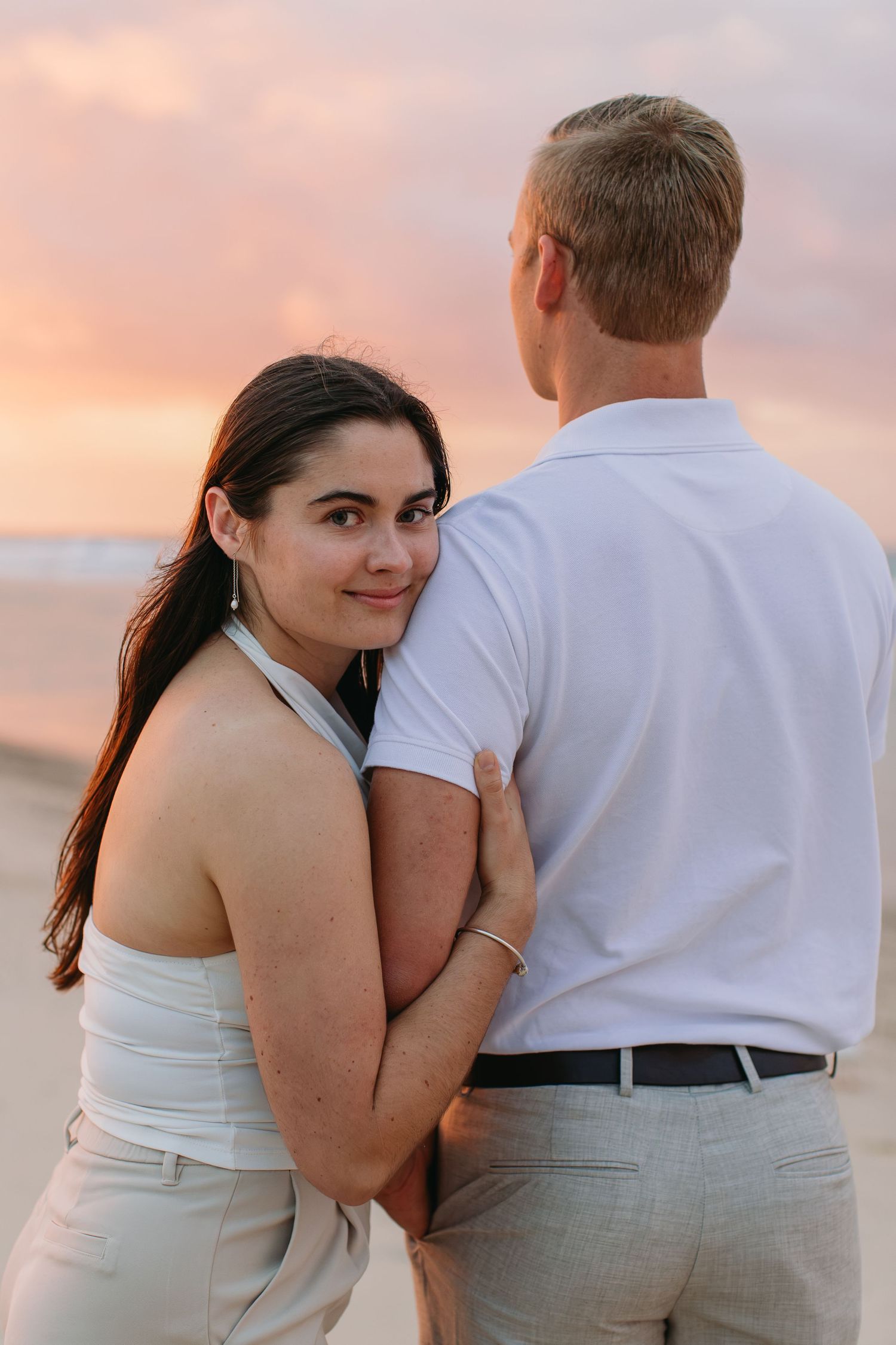 Couple embracing on beach at sunset, woman in white top looks back smiling, man in white shirt, pink sky backdrop.